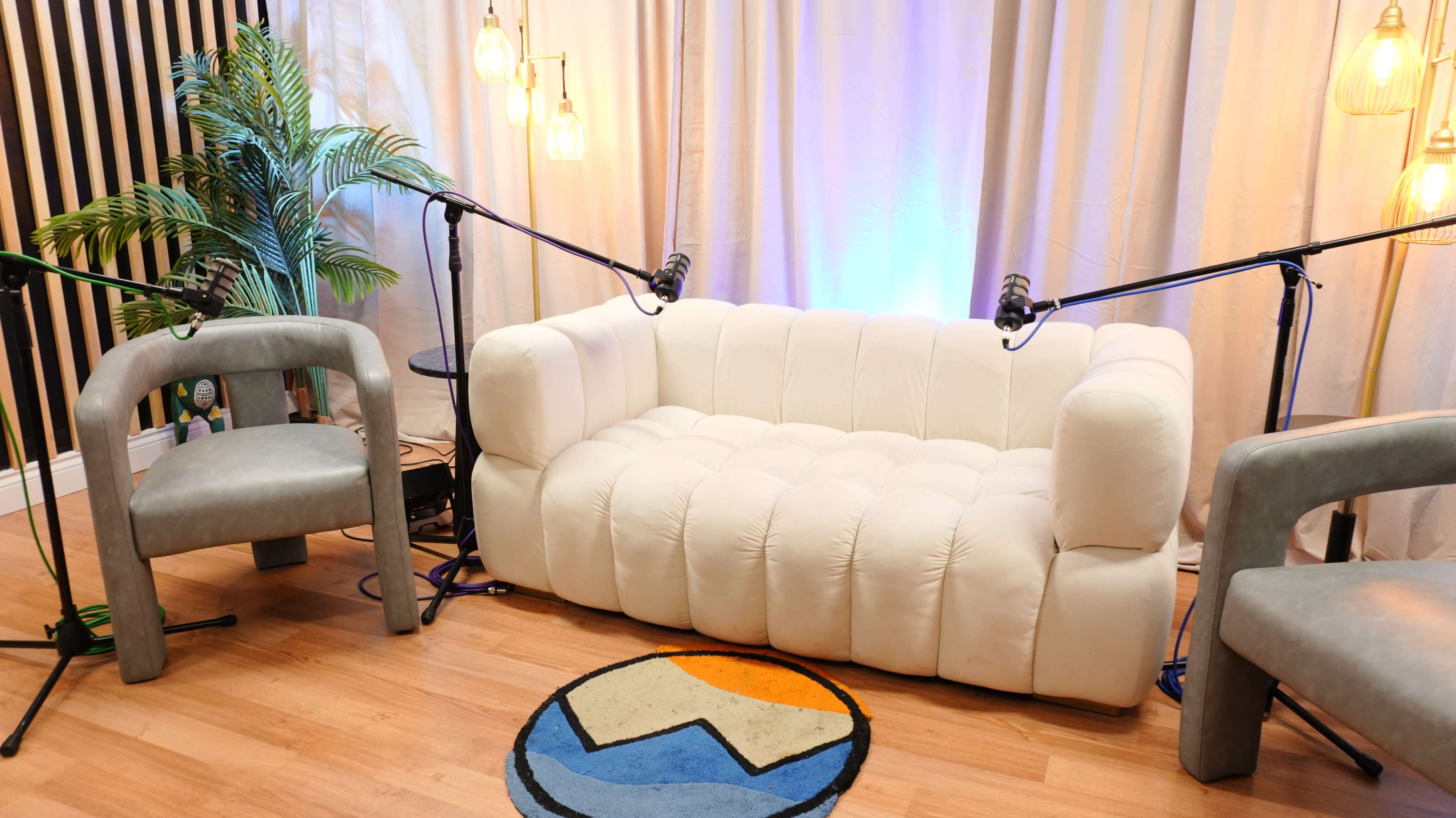 The image shows a cozy podcast recording setup featuring a white tufted sofa and two gray armchairs, with microphones positioned for speakers and soft lighting in the backdrop.