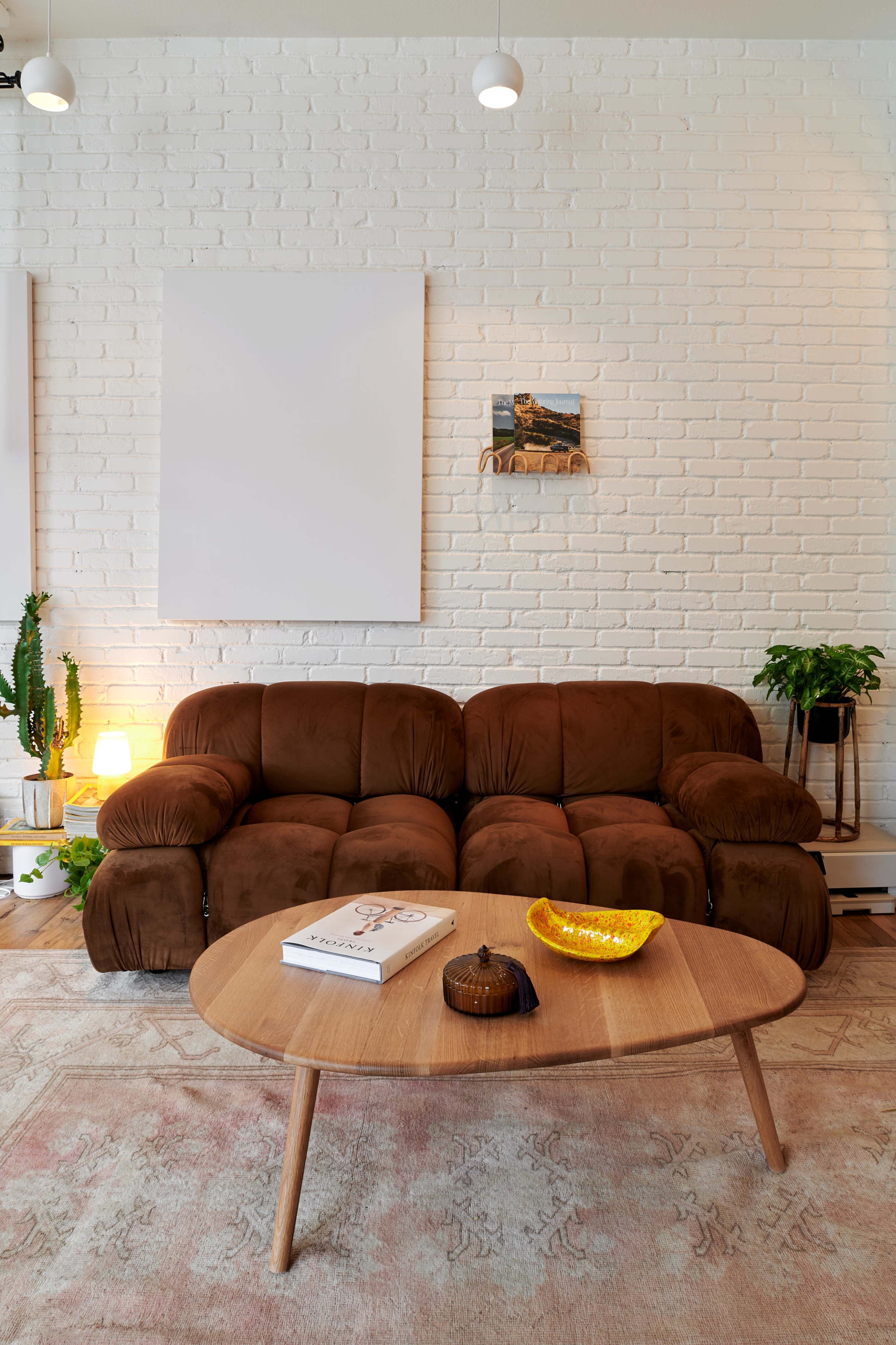 A cozy living room with a brown sofa, a round wooden coffee table, and various plants against a white brick wall.