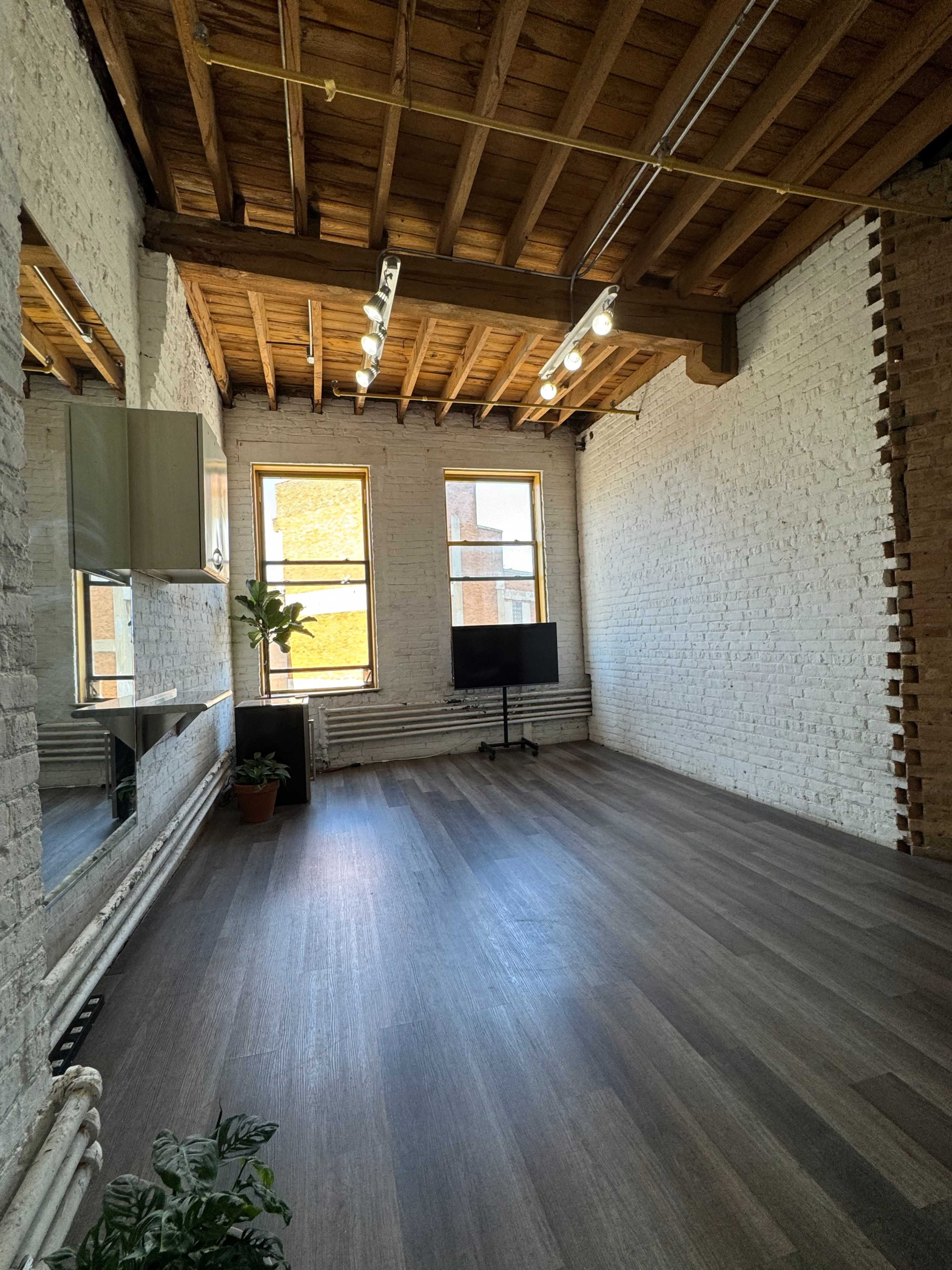 The image shows an empty room with exposed brick walls, large windows, wooden beams, and a mirror along one wall.