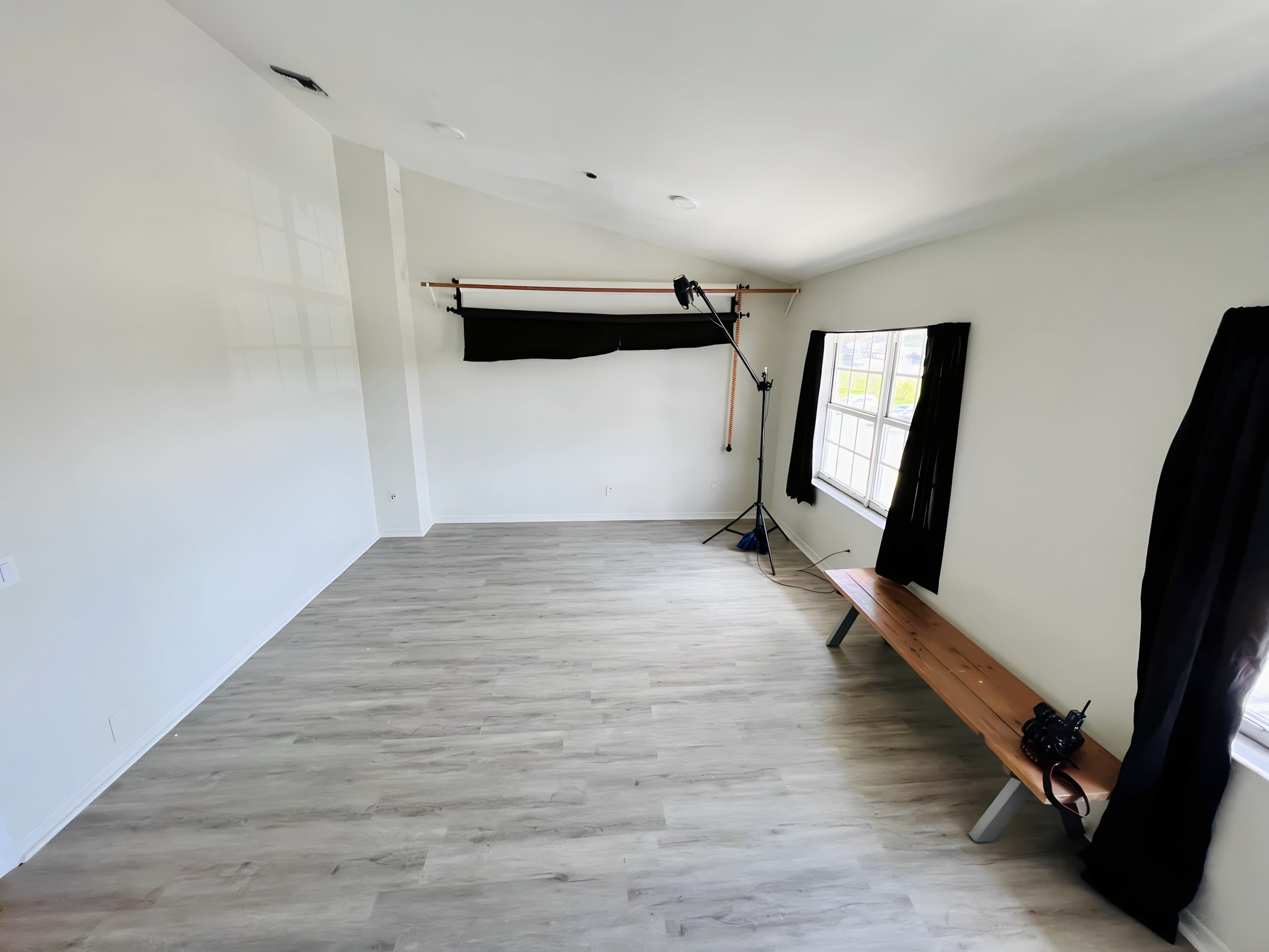 An empty room with light wood flooring, a bench along one wall, and a window with black curtains.