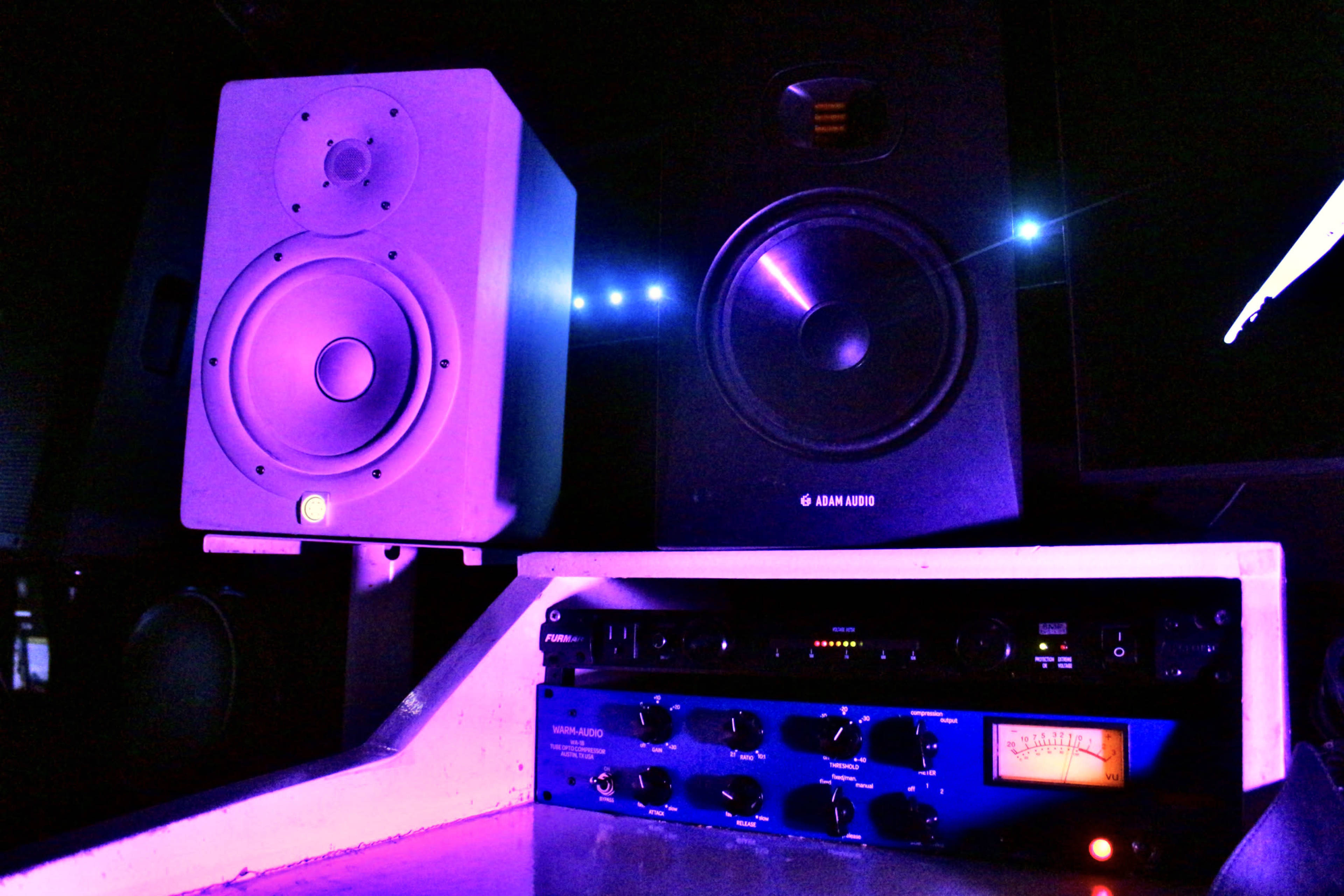 The image shows two speaker monitors and an audio interface set against a dark background with purple lighting.