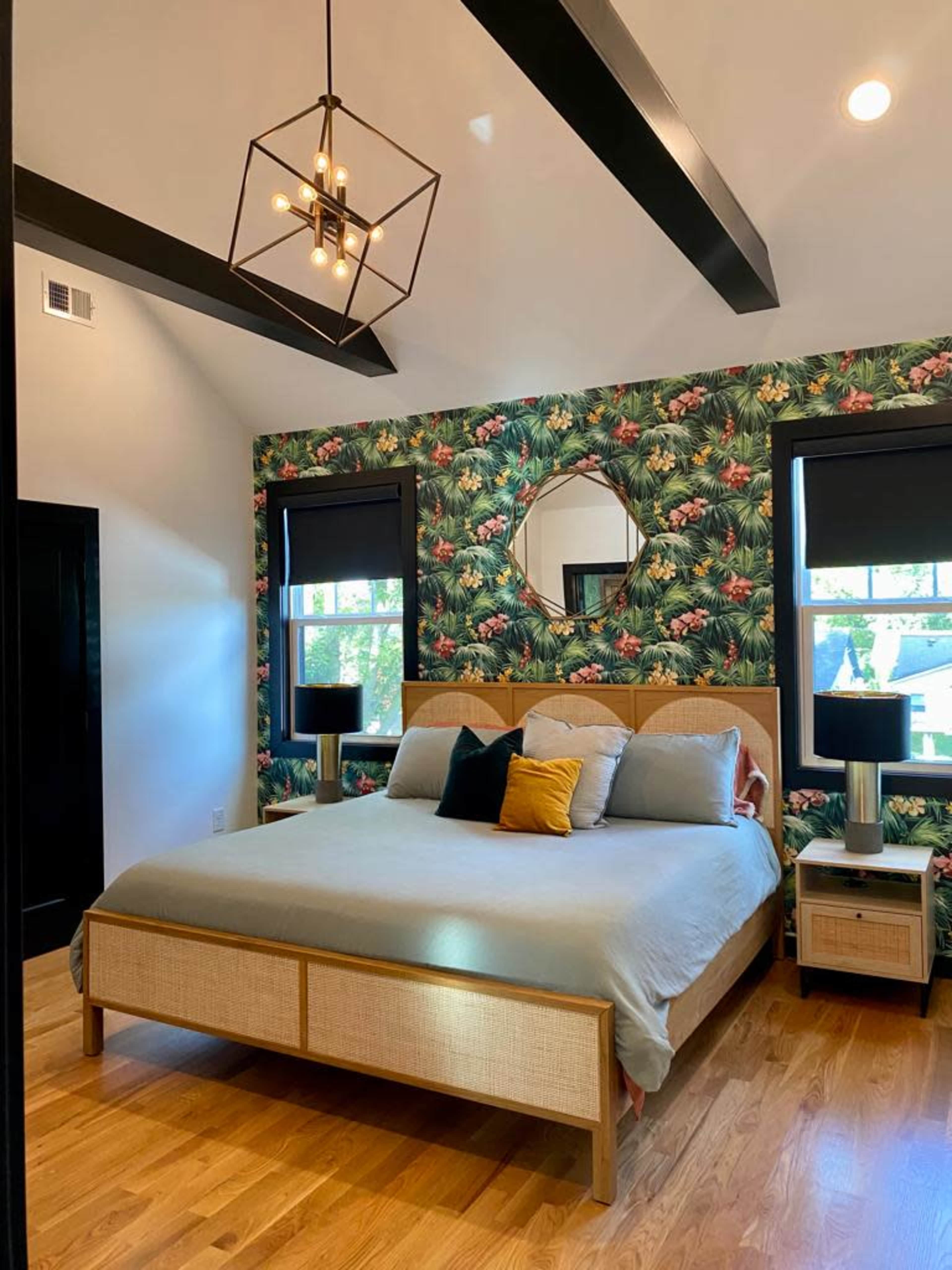 The bedroom features a large bed with neutral bedding, surrounded by floral wallpaper, two lamps on nightstands, and a round mirror above the headboard.