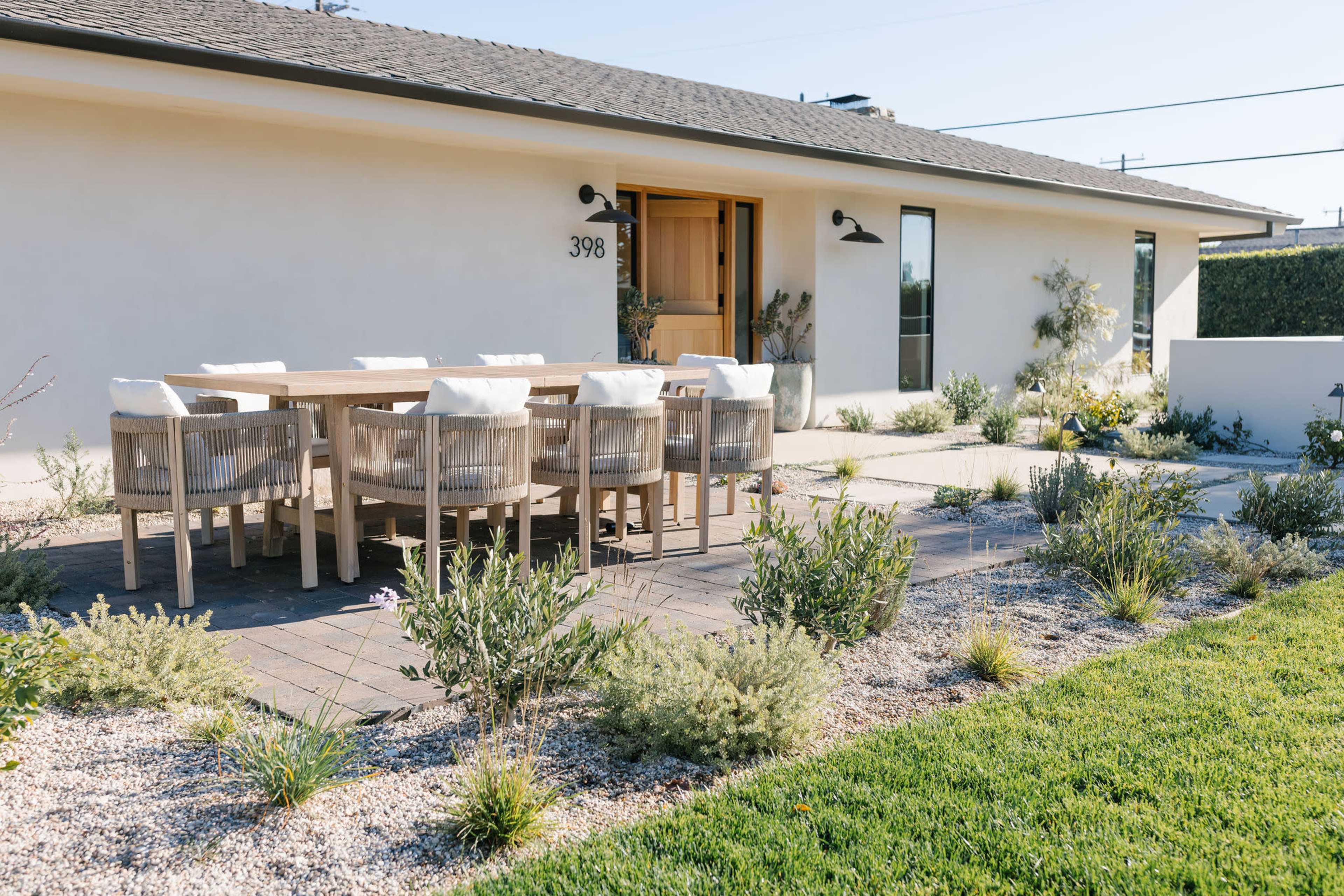 A patio with a wooden dining table and chairs is situated in front of a light-colored house surrounded by low shrubs and grass.