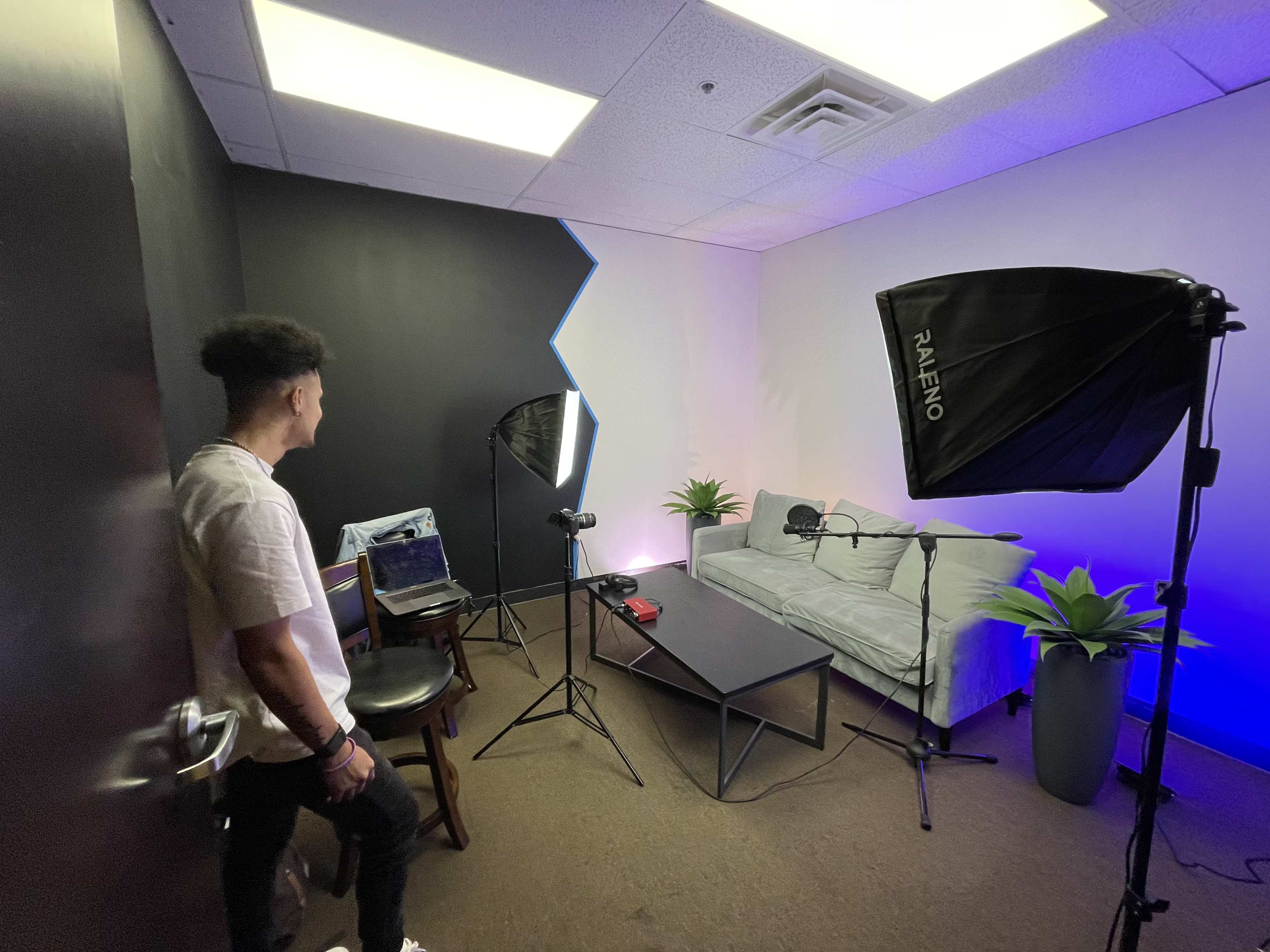A person stands near a setup for a video recording in a small room with lighting equipment, a microphone, and a sofa.