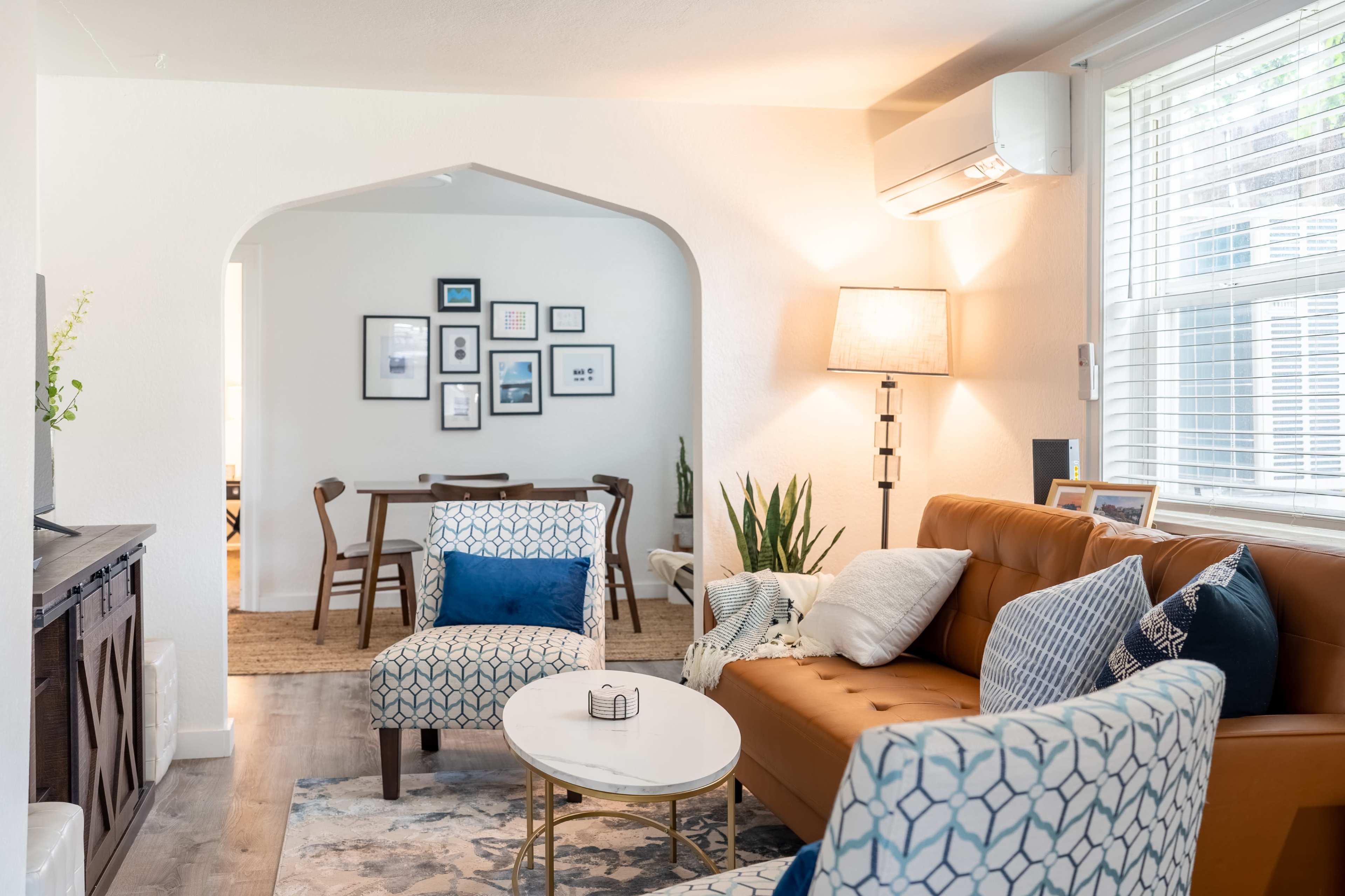 A living room with a brown leather couch, two patterned chairs, a round coffee table, and an archway leading to a dining area.