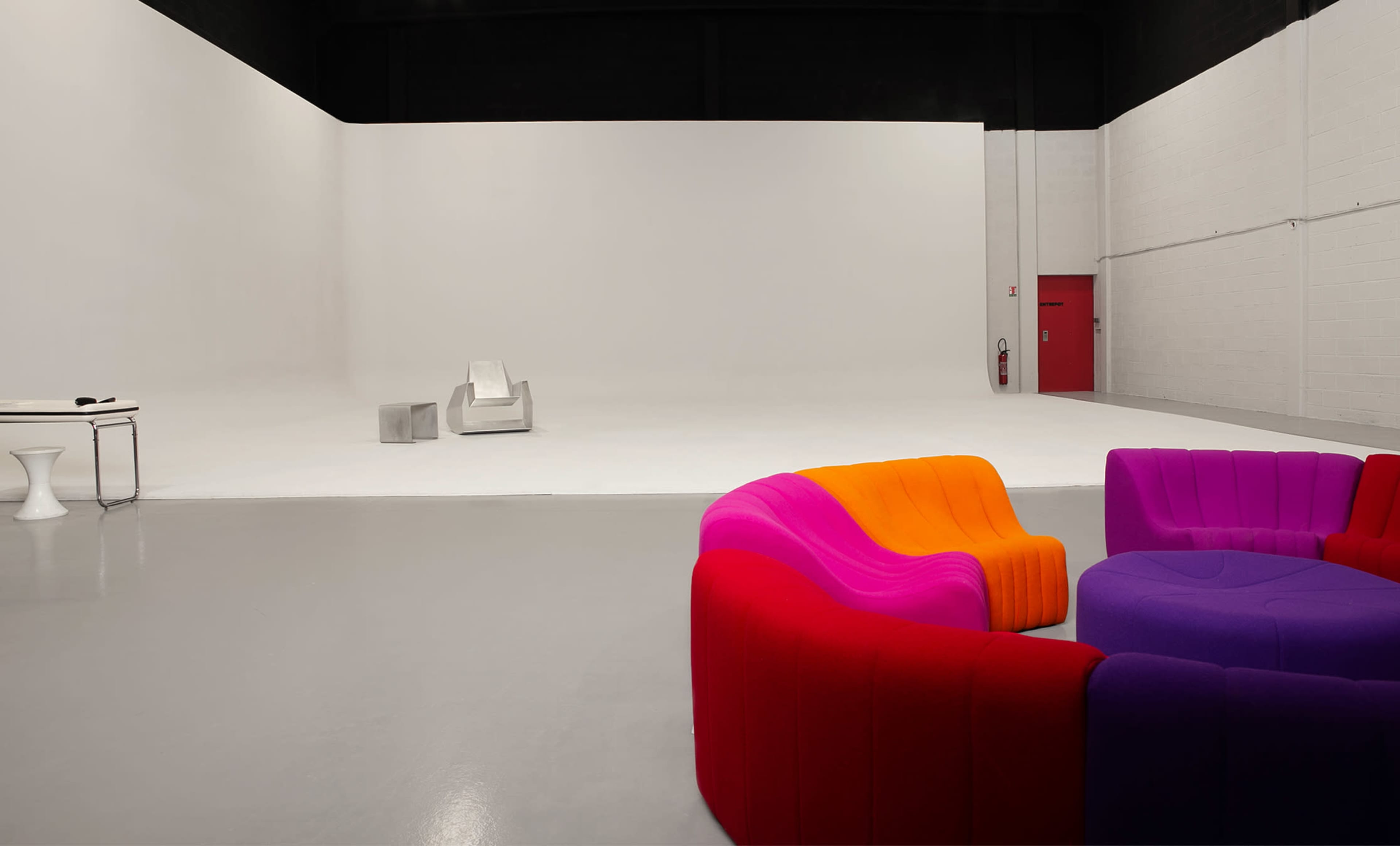 The image shows a spacious, minimalist studio with a white backdrop, featuring colorful seating in the foreground and a couple of chairs in the center.