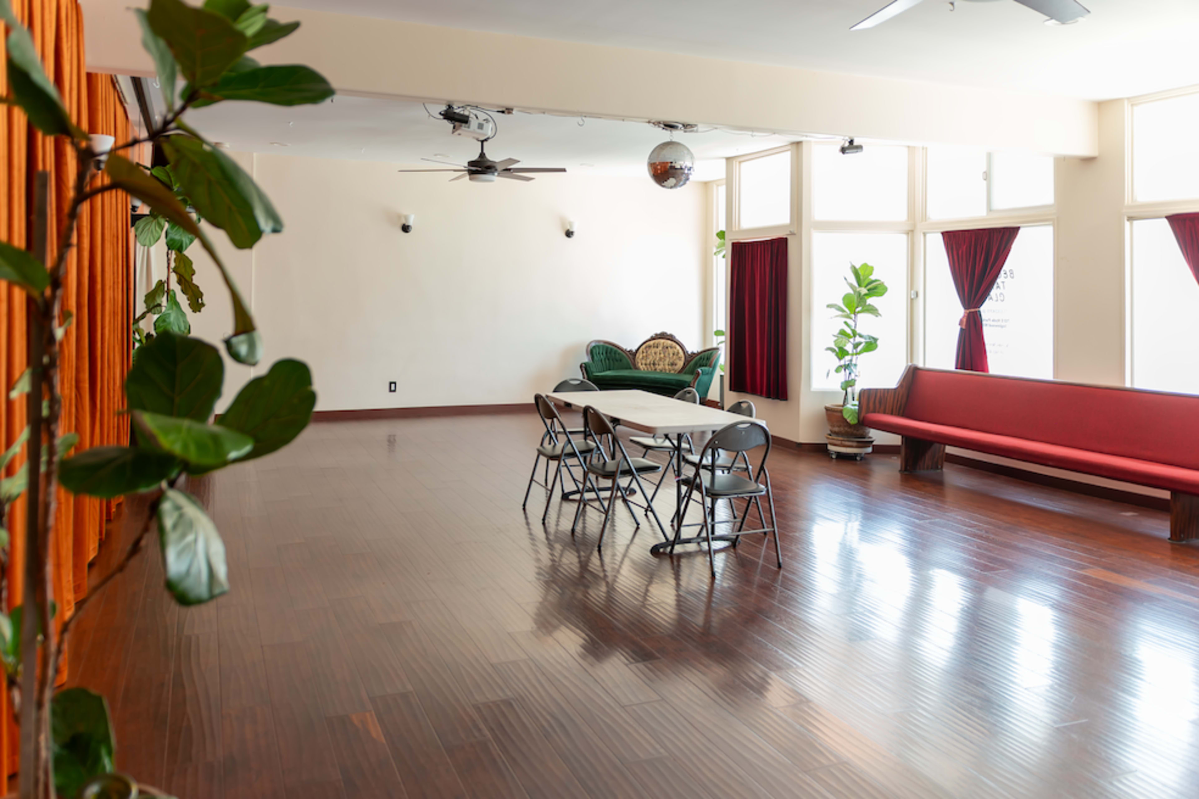 The image shows a spacious room with hardwood floors, featuring a long table with chairs, a red couch, potted plants, and large windows with burgundy curtains.