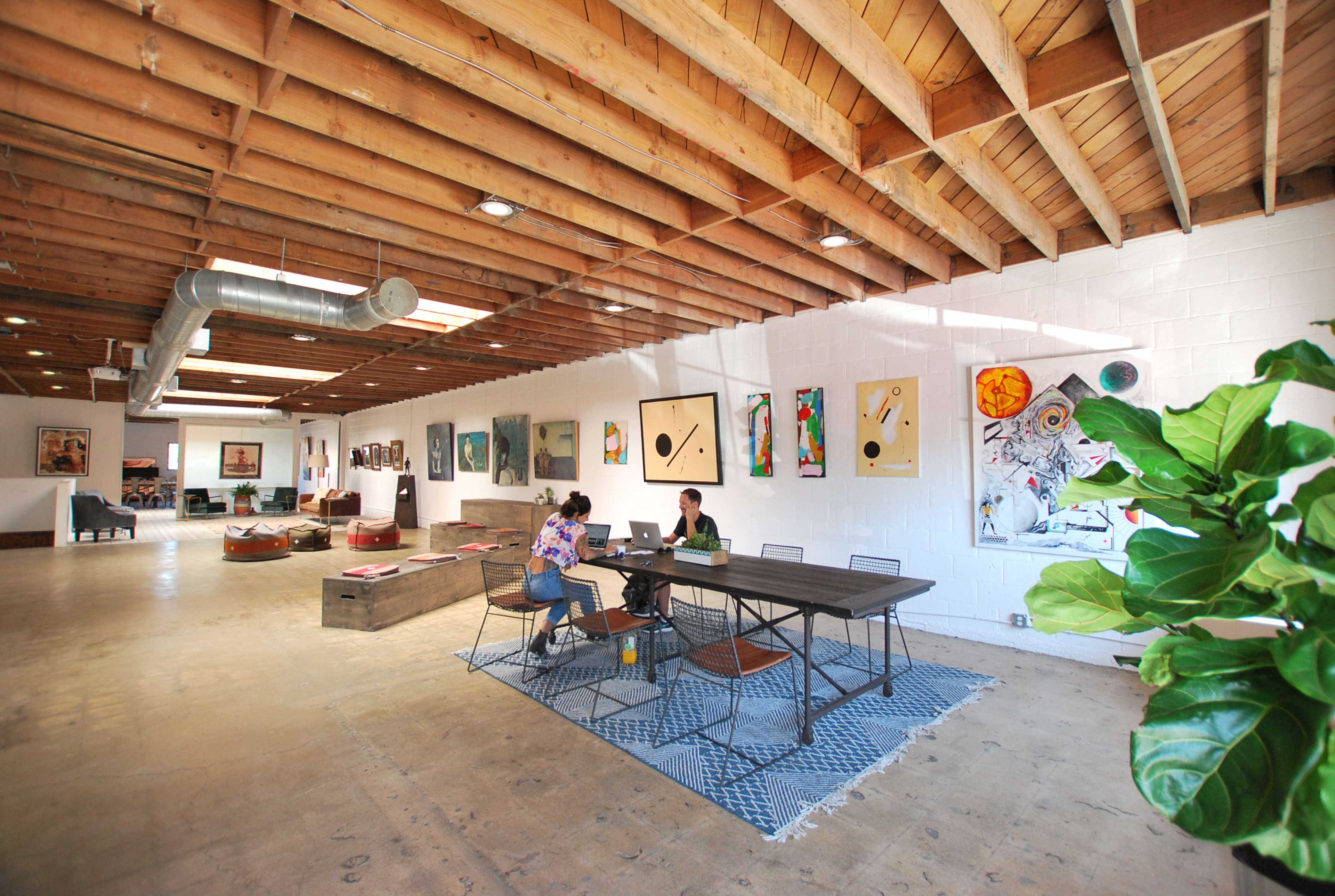 Two people are working at a table in a spacious art studio with wooden beams and various artworks displayed on the walls.