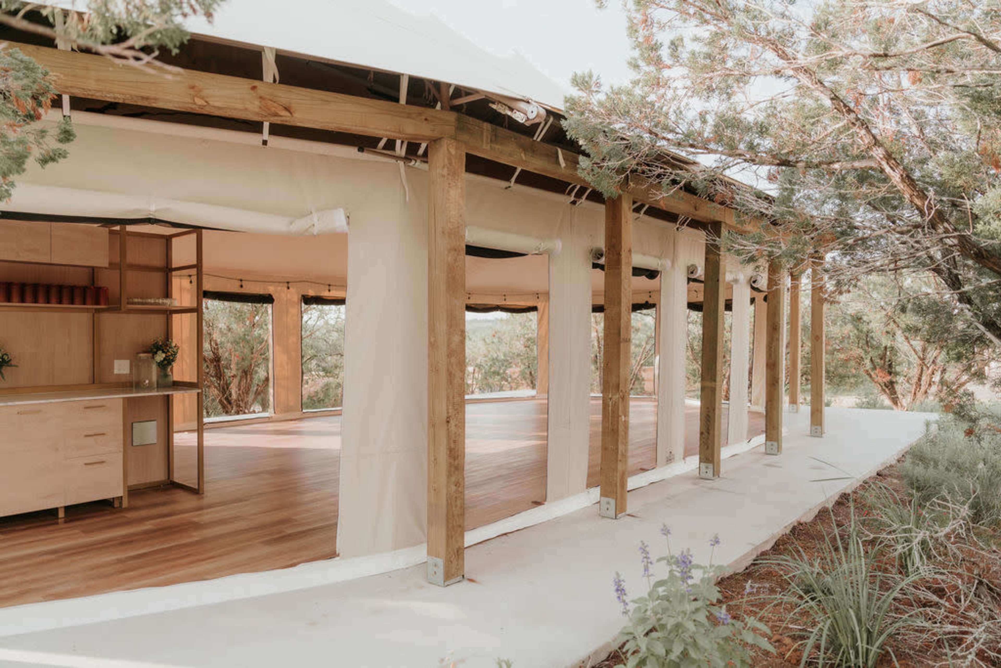 A modern, partially open structure with wooden beams and large fabric walls sits among greenery, featuring a spacious wooden deck and interior cabinetry.