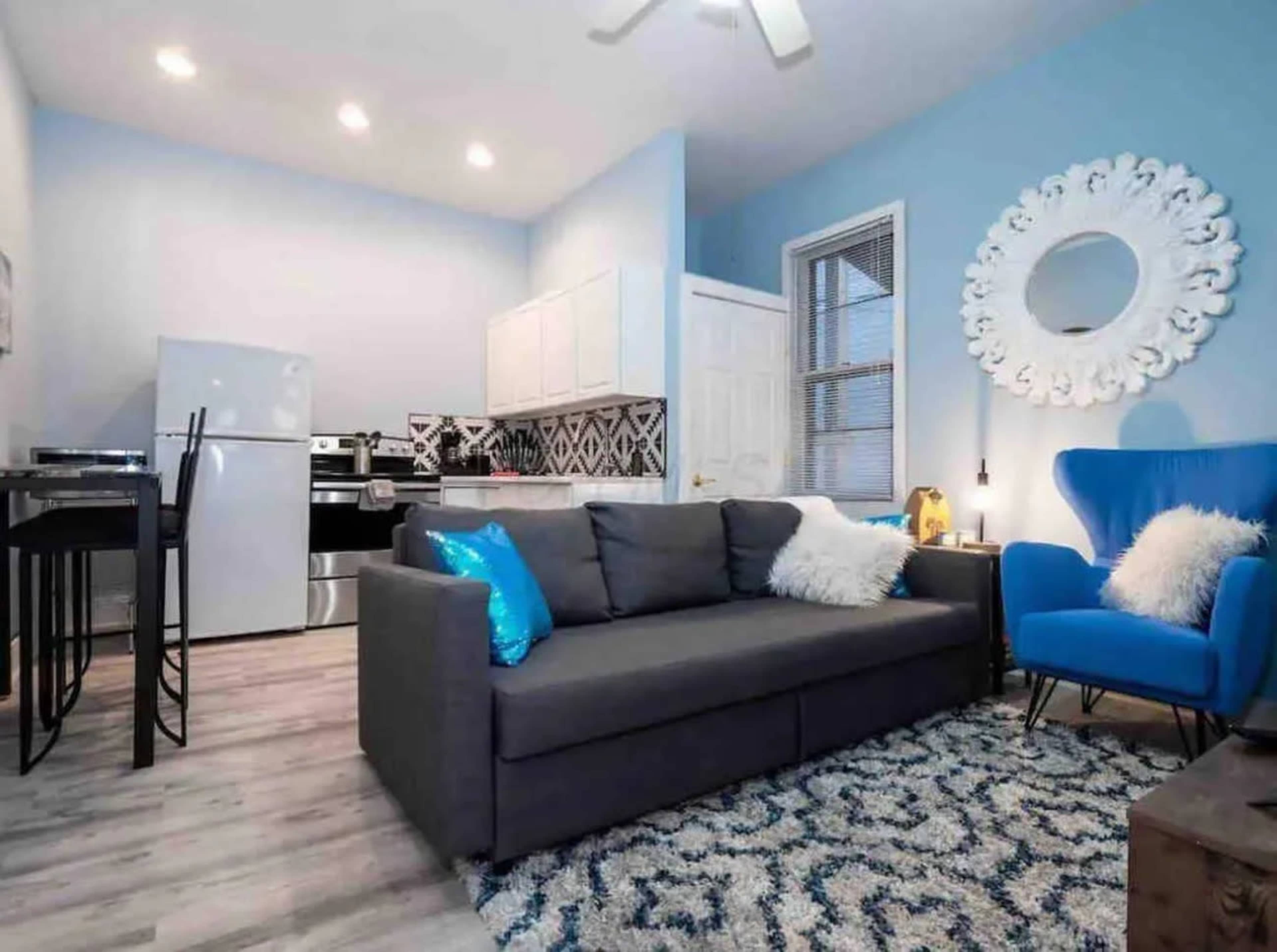 The image shows a small, modern living area featuring a gray sofa, a blue accent chair, a dining table, and a kitchen area with white cabinets and stainless steel appliances.