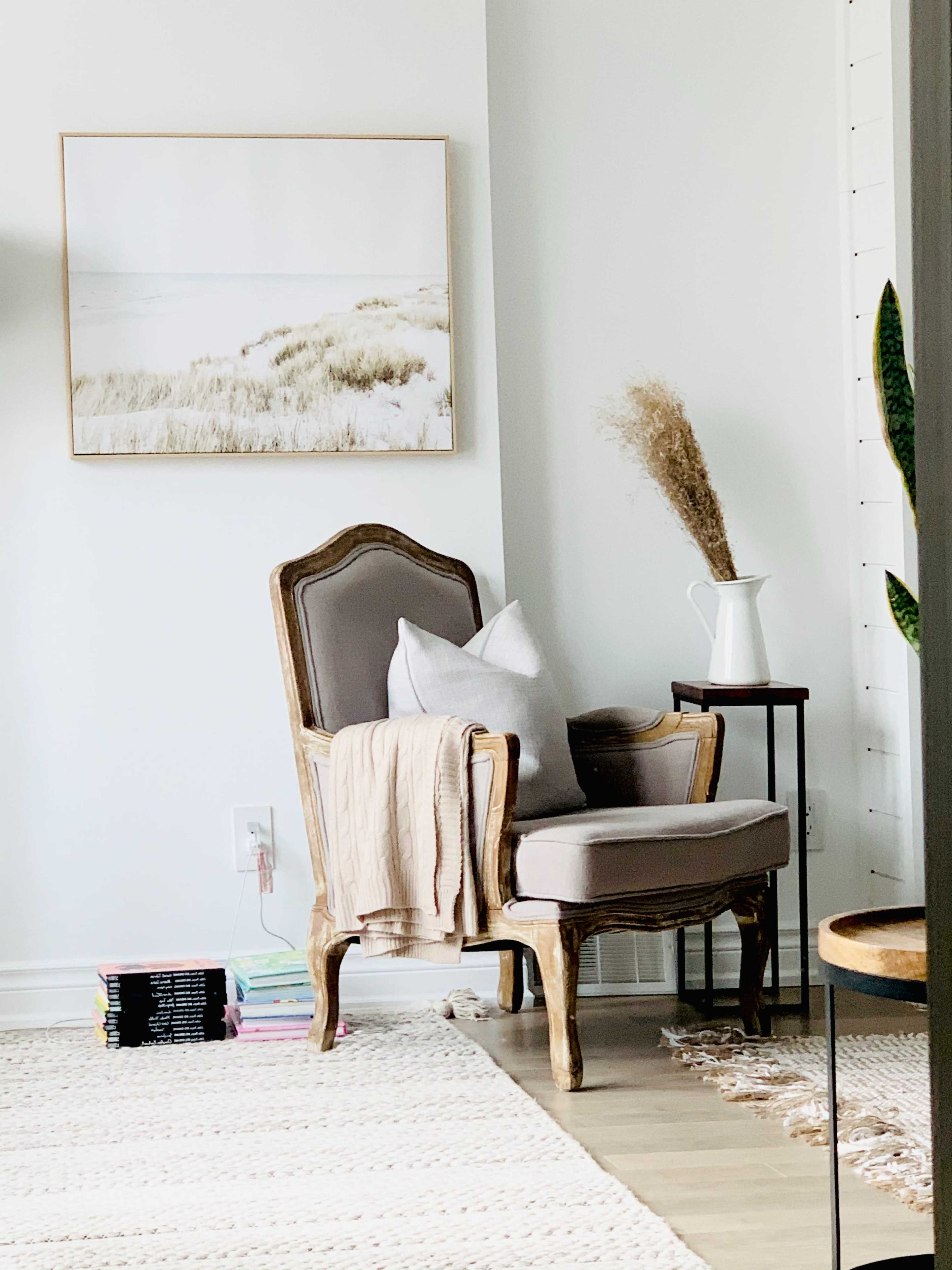 A vintage armchair with a throw blanket sits beside a small side table and a decorative vase in a bright, minimalistic room.