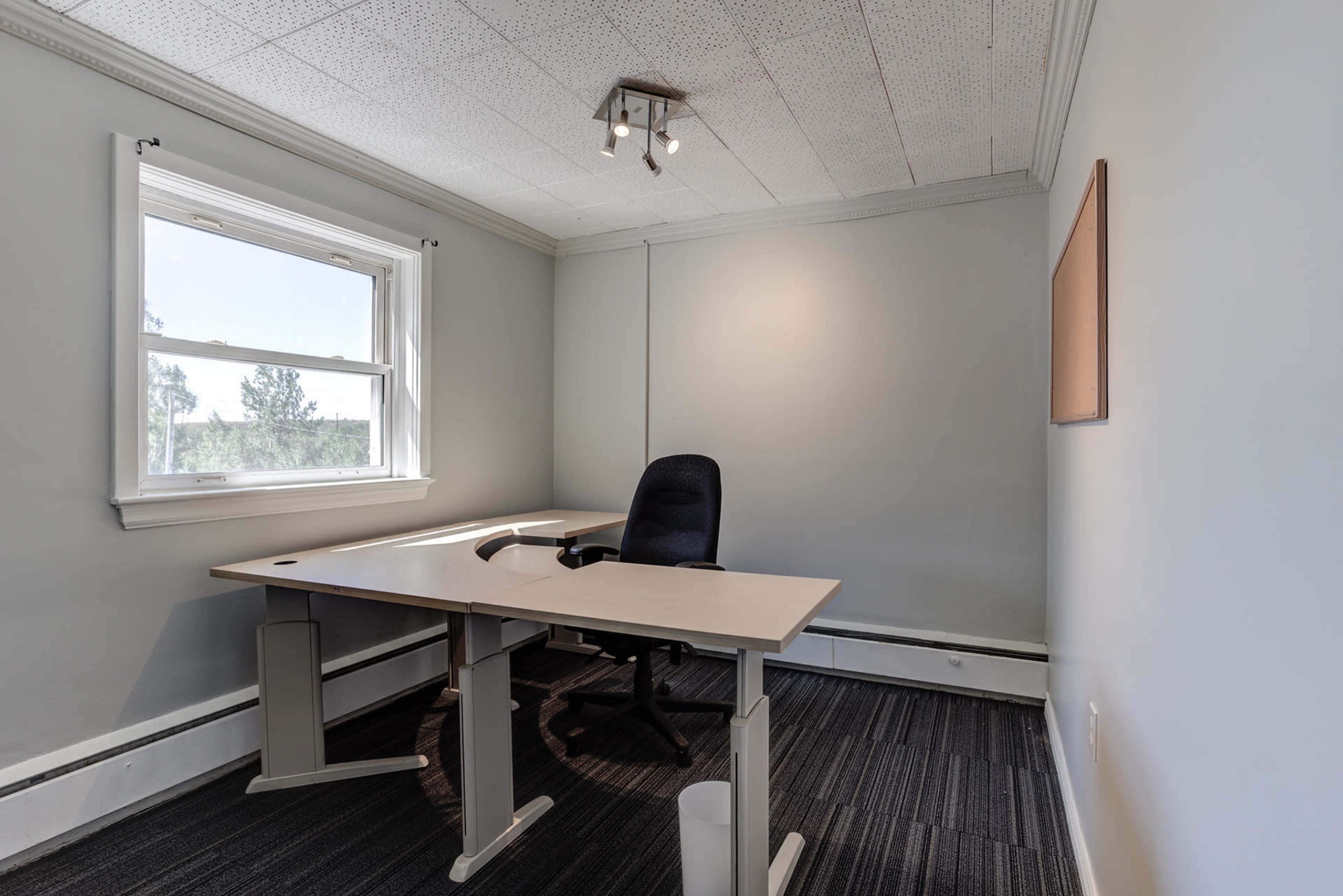 A dimly lit office features a single desk and chair positioned against a gray wall, with a window letting in natural light.