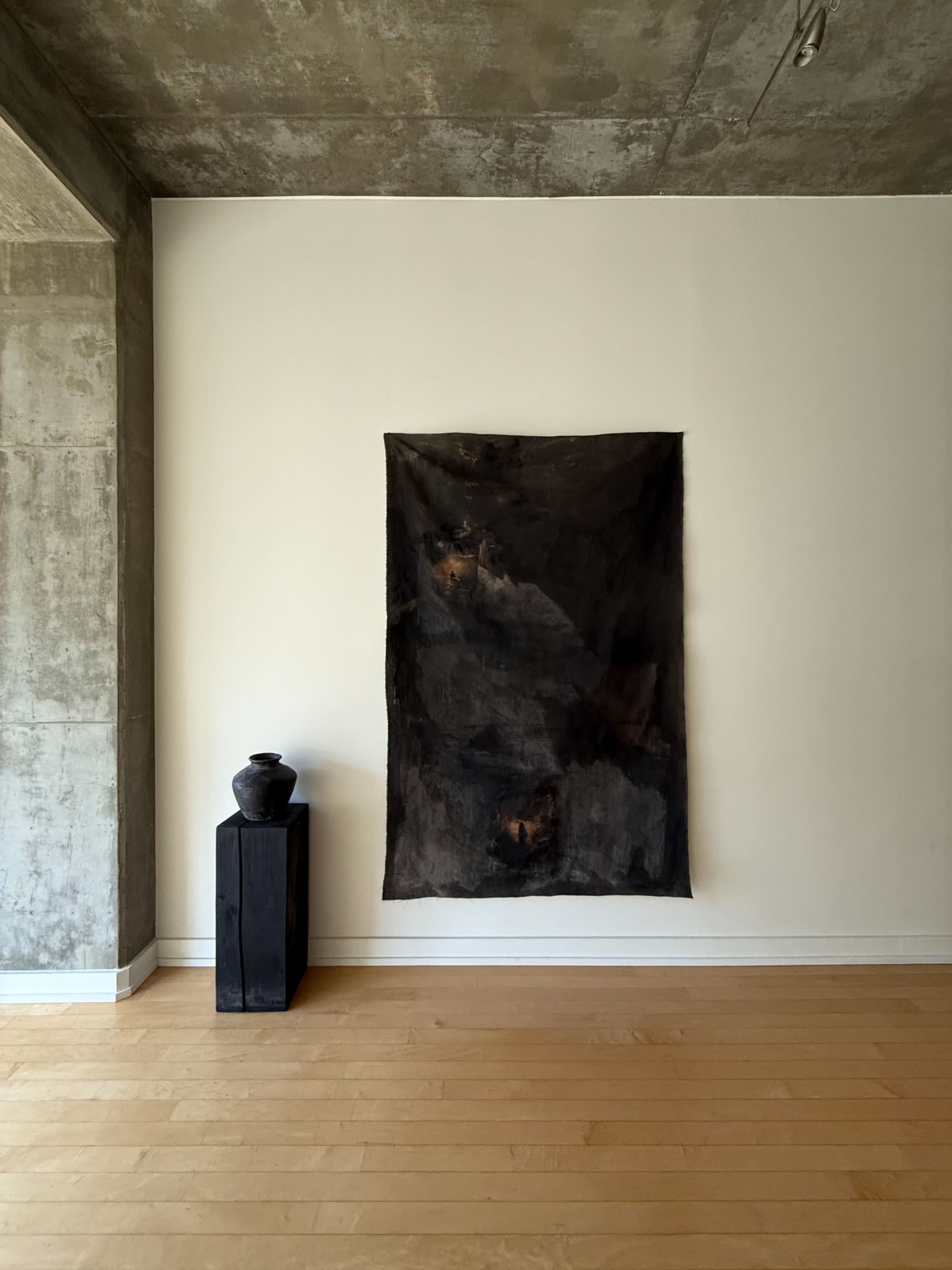 A dark textile is hung on a white wall above a black wooden pedestal with a ceramic vase on top, set against a backdrop of a concrete wall and wooden floor.