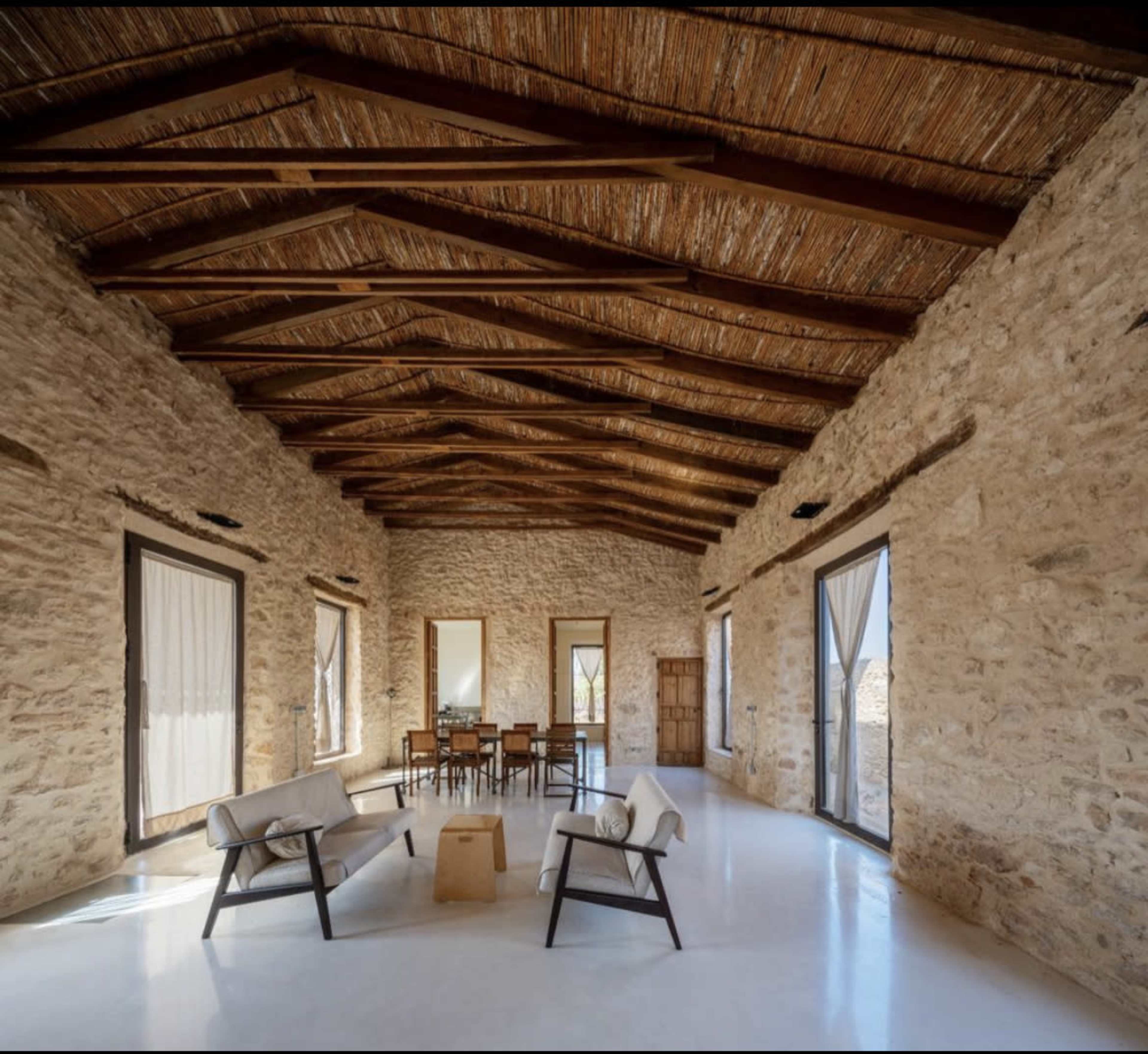 The image shows a spacious interior of a stone-walled room with wooden beams, featuring a seating area and a dining table.