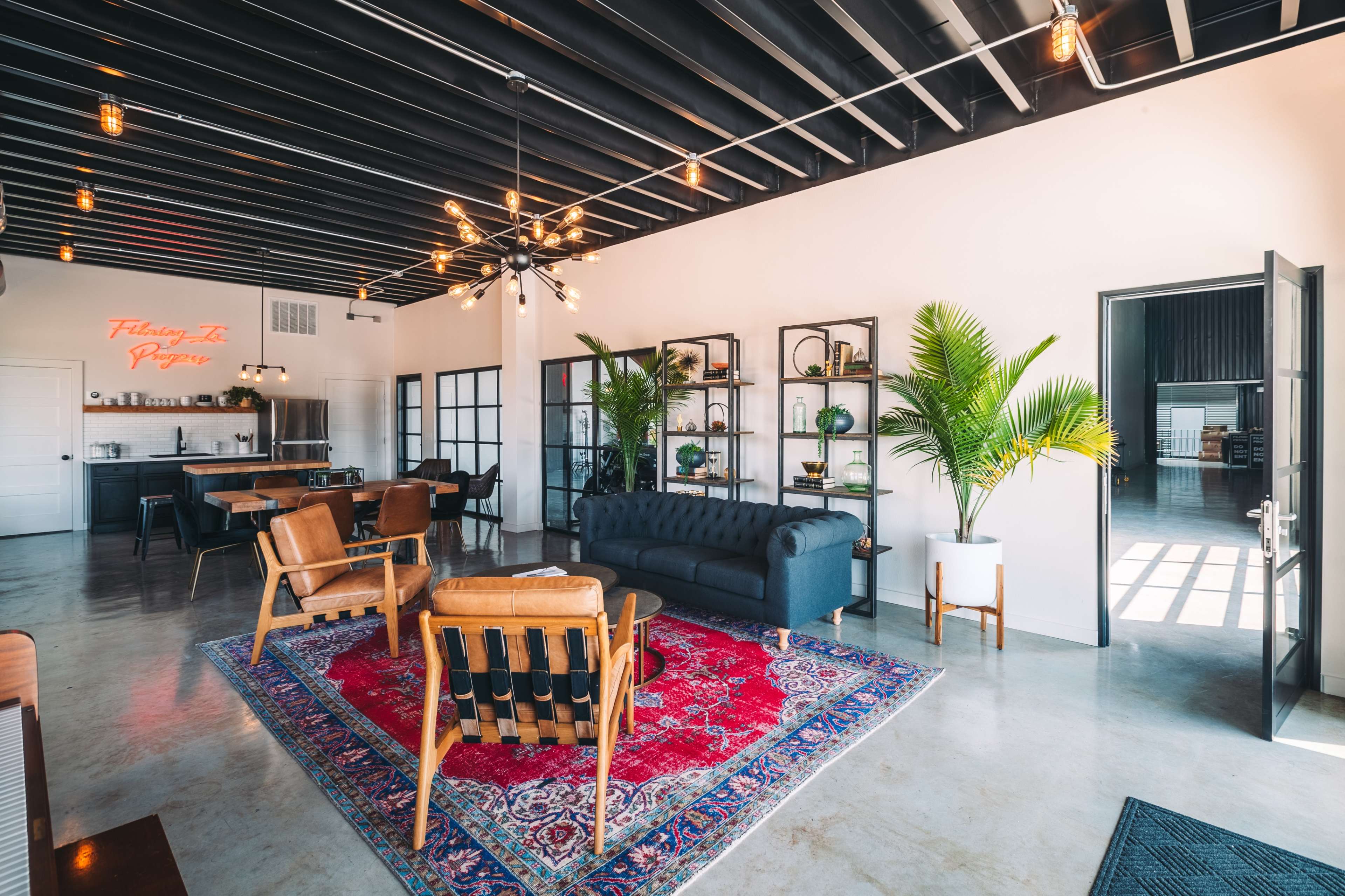 The image shows a modern lounge area with a large black sofa, wooden chairs, a red rug, and shelves filled with plants, illuminated by a decorative light fixture and neon signage.