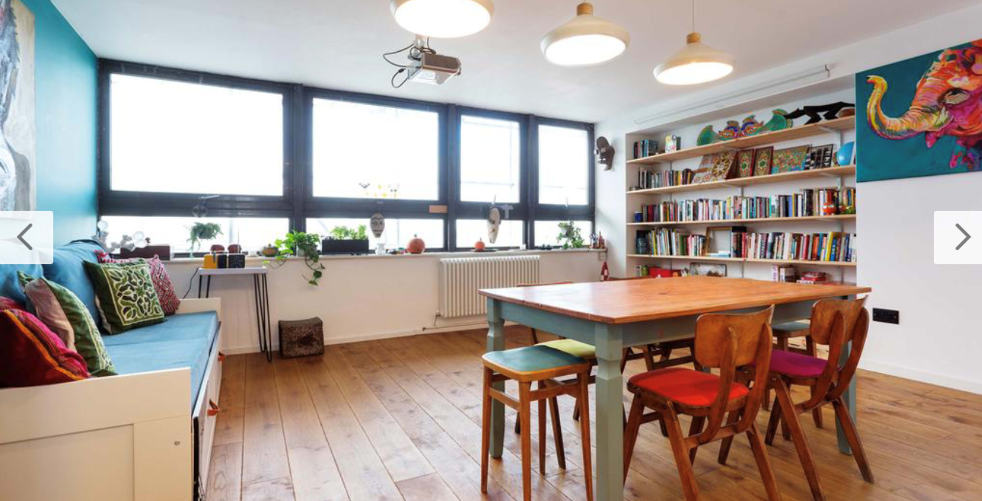 A bright, modern living room with a wooden dining table surrounded by colorful chairs, a large bookshelf filled with books, and an elephant painting on the wall.