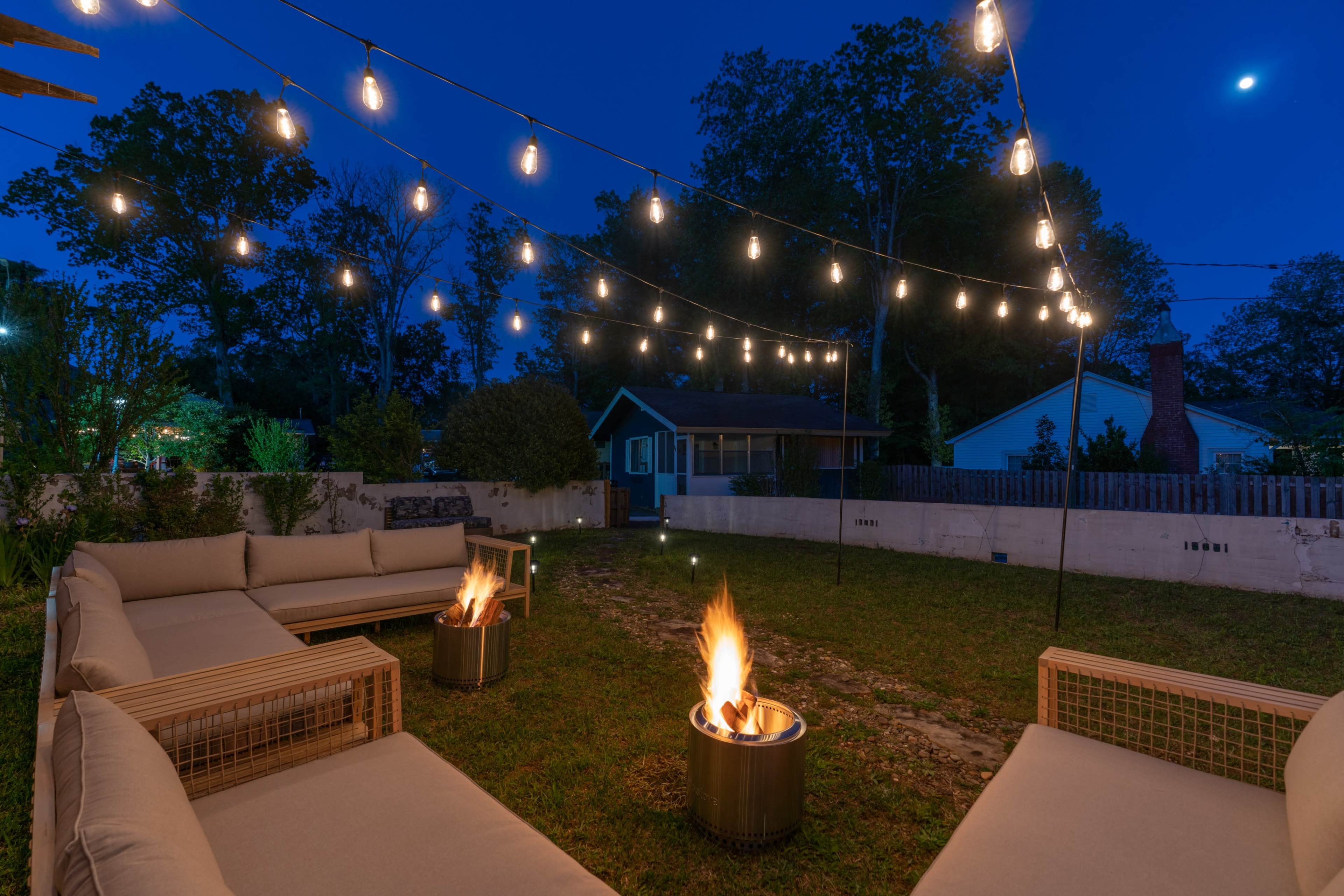 A cozy outdoor seating area with fire pits and string lights illuminates a grassy space at dusk.