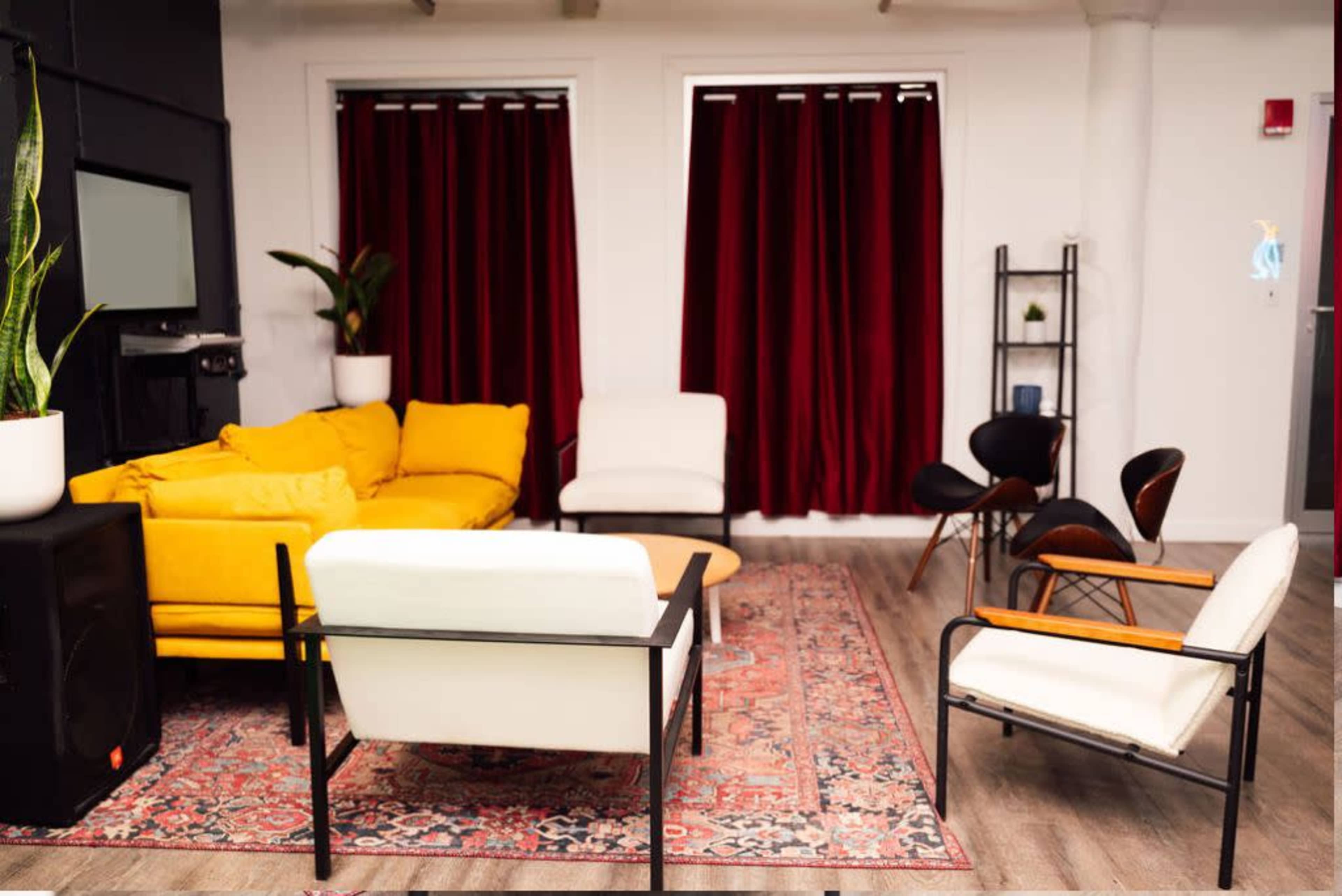 A modern living room features a yellow sofa, two white chairs, a wooden coffee table, a patterned rug, and red curtains beside a window.