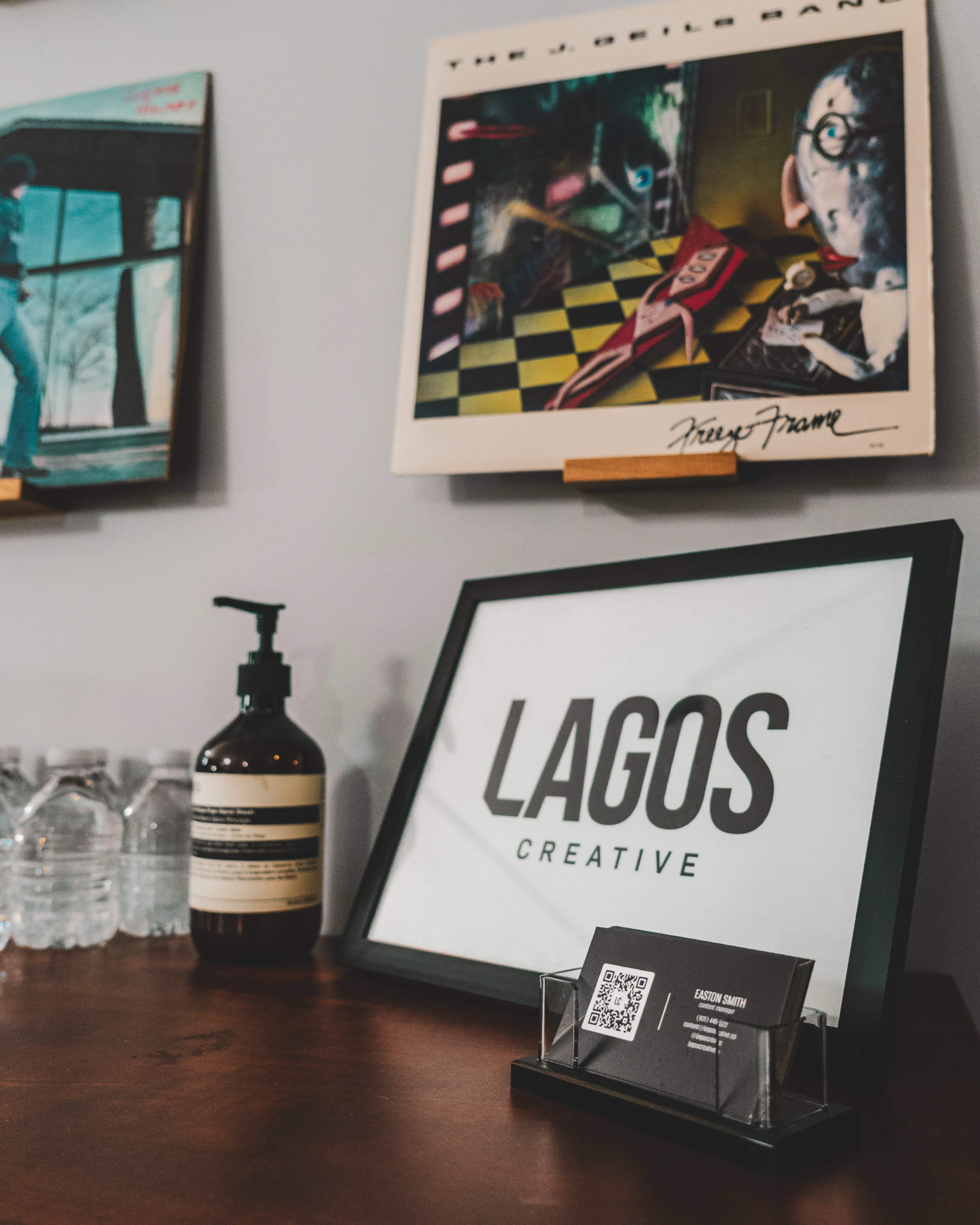 The image shows a wooden table displaying a framed sign that reads "LAGOS CREATIVE," a business card with a QR code, a bottle of hand sanitizer, and several bottles of water, with vinyl records hanging on the wall behind.
