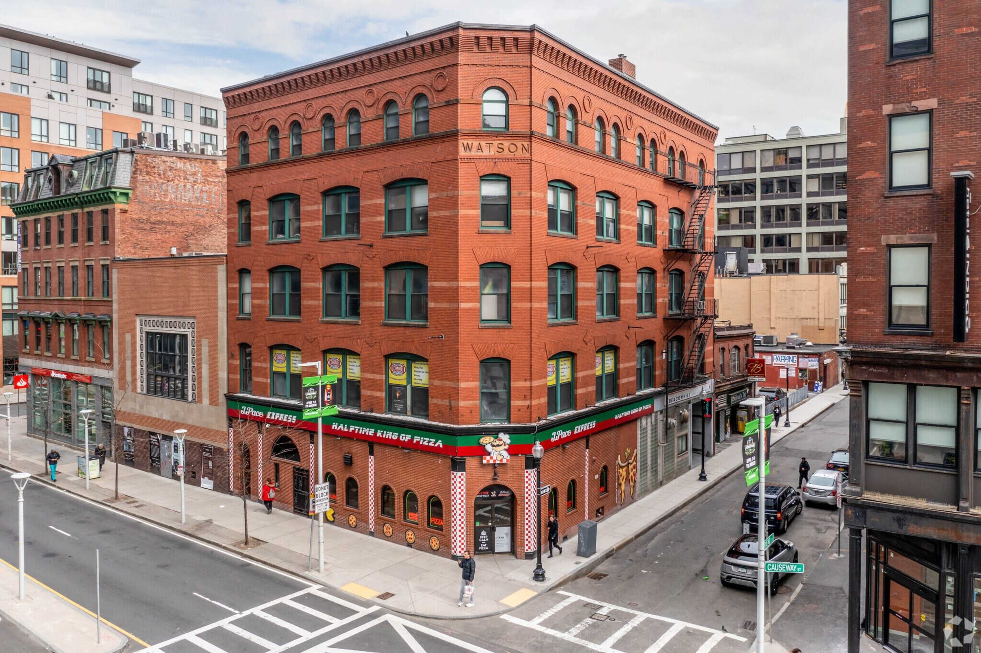 A red brick building with the name "WATSON" on its facade, located at the corner of a street with a pizza restaurant and surrounding urban structures.