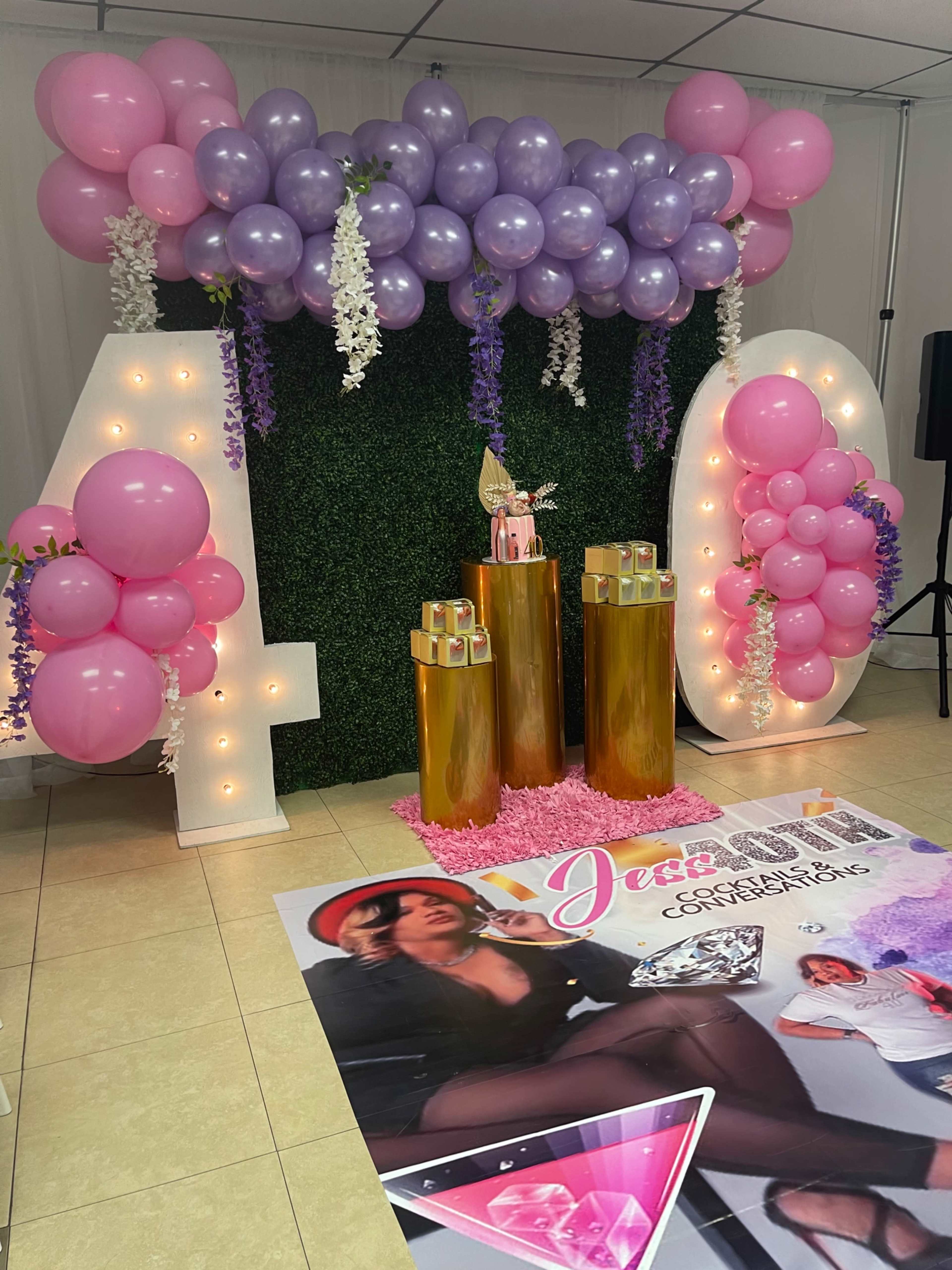 The image shows a decorated party backdrop featuring large illuminated numbers "40," colorful balloons, and a display with a promotional poster and decorative elements.