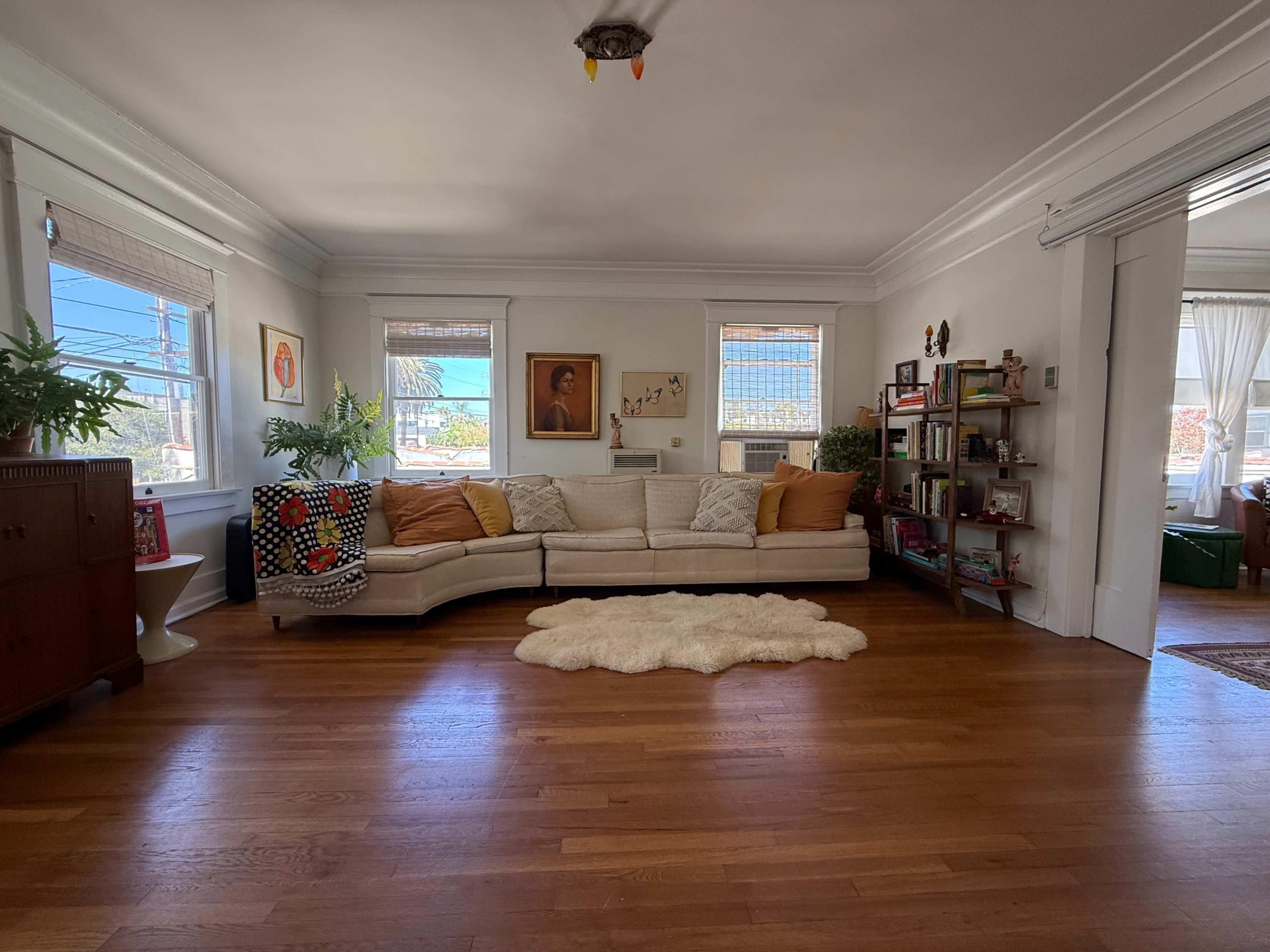 The living room features a curved white sofa with orange throw pillows facing a wooden bookshelf and a bright window.