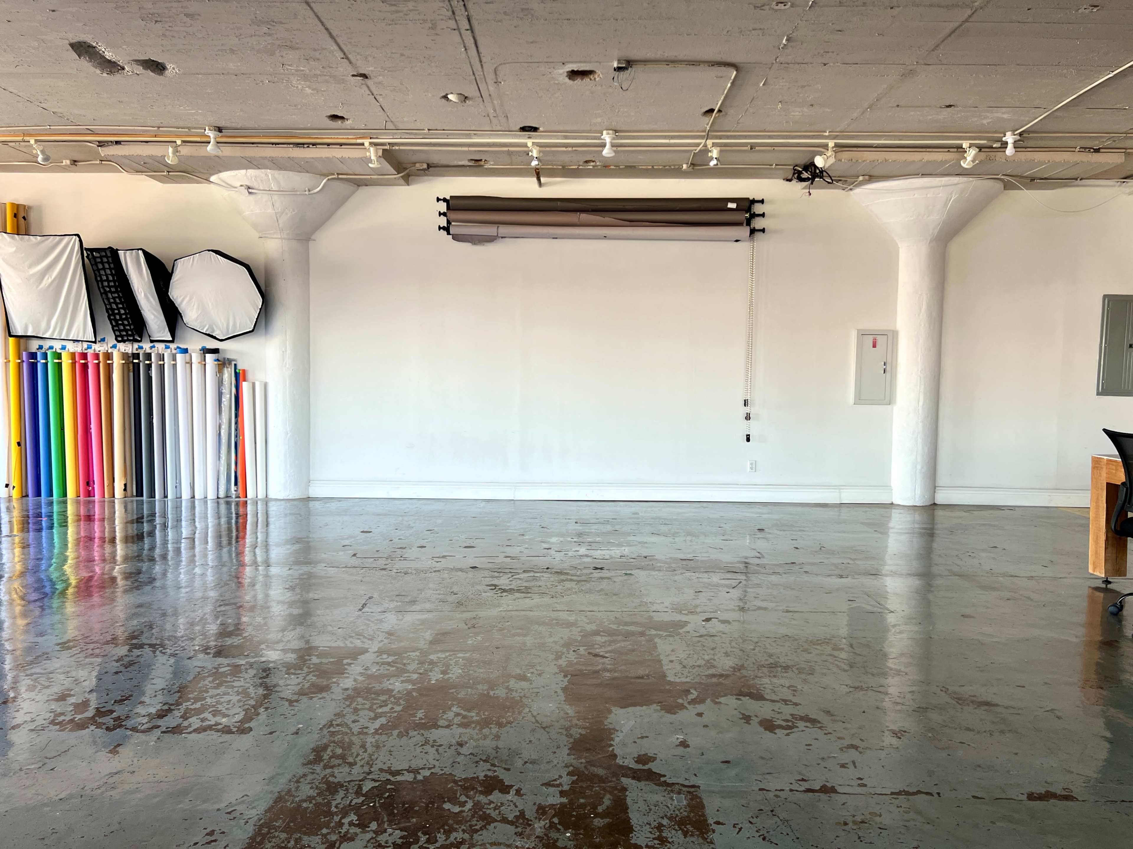 The image shows a spacious studio with white walls, polished concrete floors, and colorful backdrop rolls lined against one side.