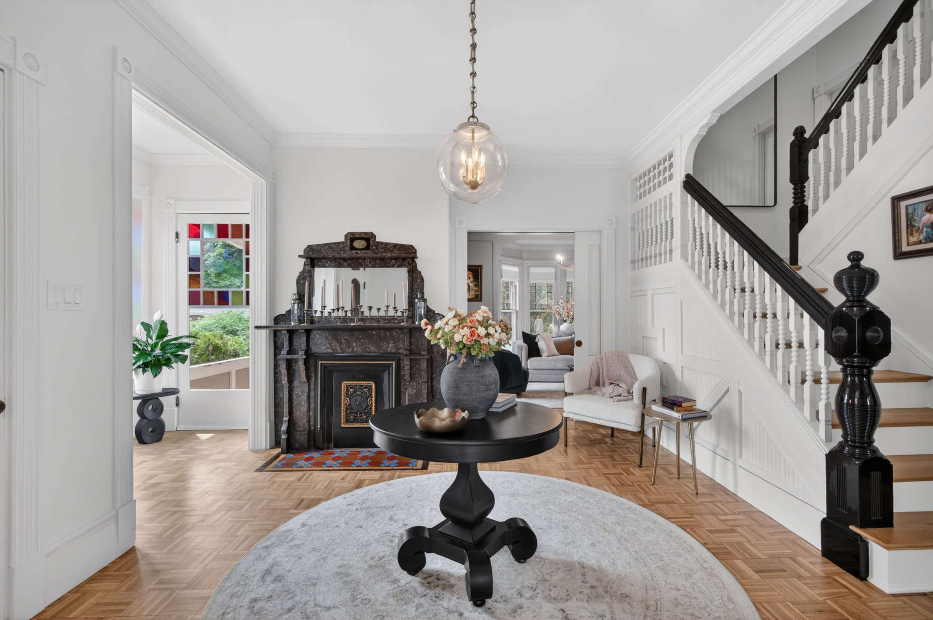 The image shows a bright entryway featuring a round black table with a floral arrangement, a staircase with a decorative banister, and a view into a living area with a fireplace and seating.
