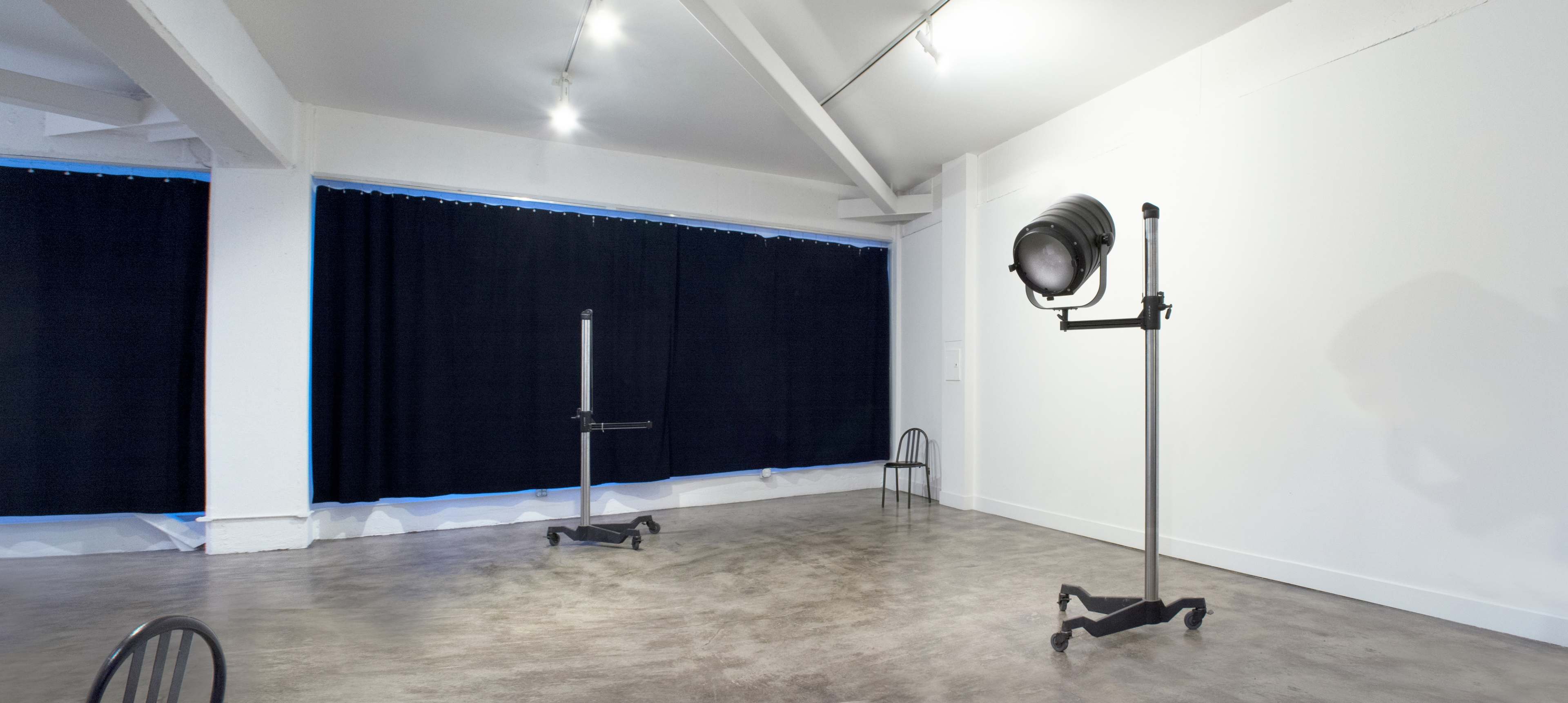 A minimalist gallery space with a concrete floor, featuring two stands with attached lighting fixtures positioned opposite each other against a backdrop of dark curtains.