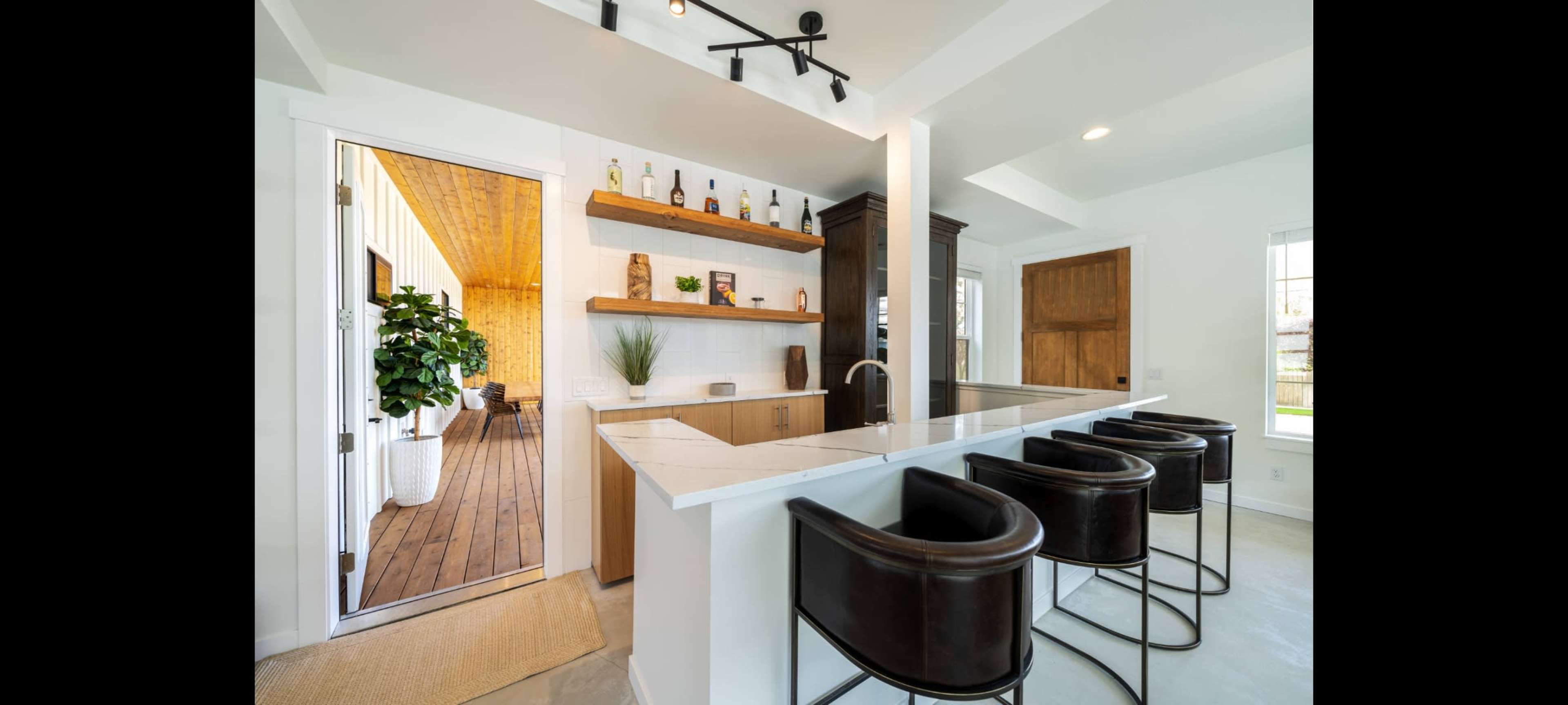A modern kitchen with a bar area featuring four black stools, open shelving with decorative items, and a doorway leading to another room.