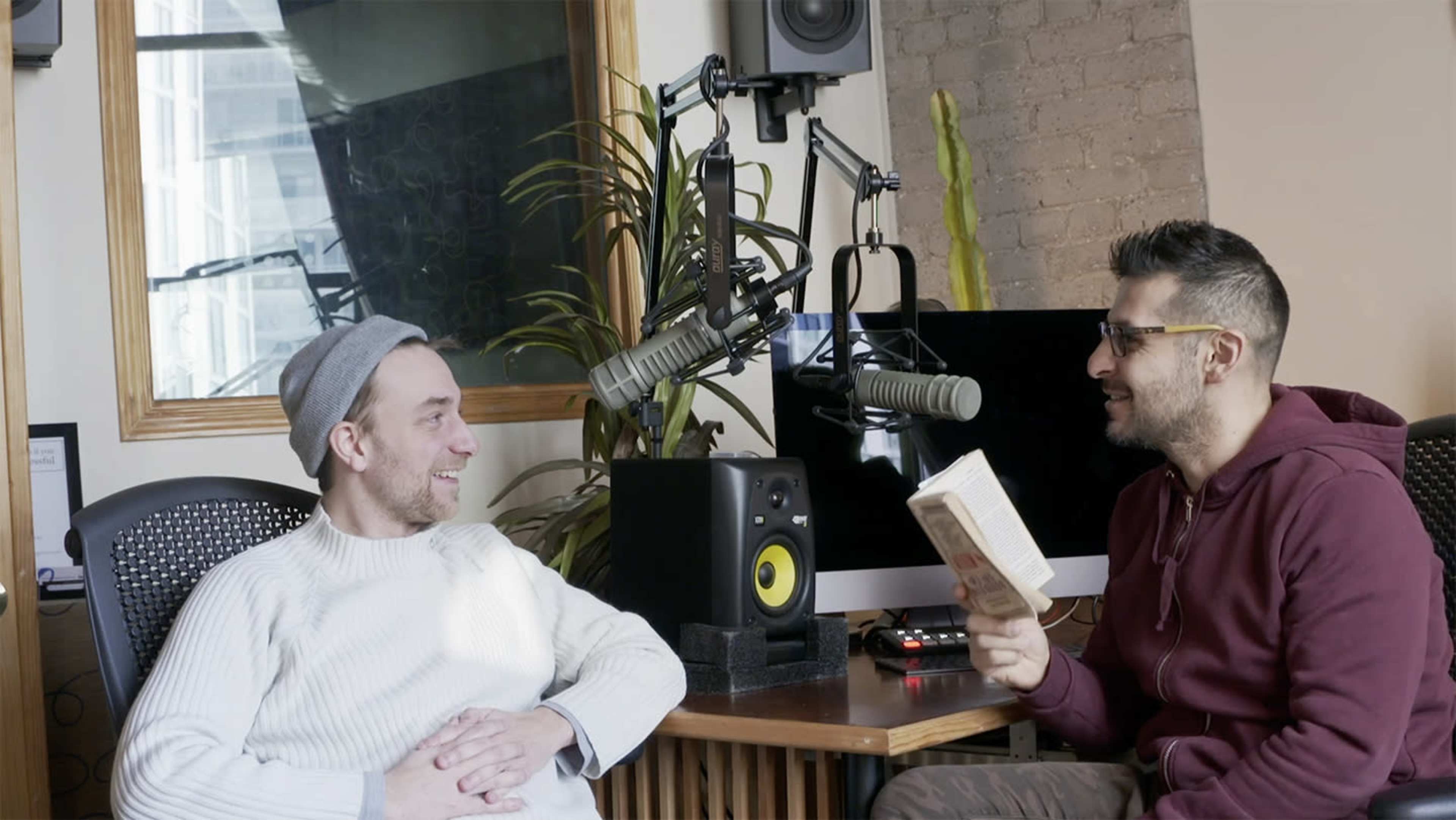 Two men sit in a studio setting, one reading from a book while the other listens, surrounded by audio equipment and a plant.