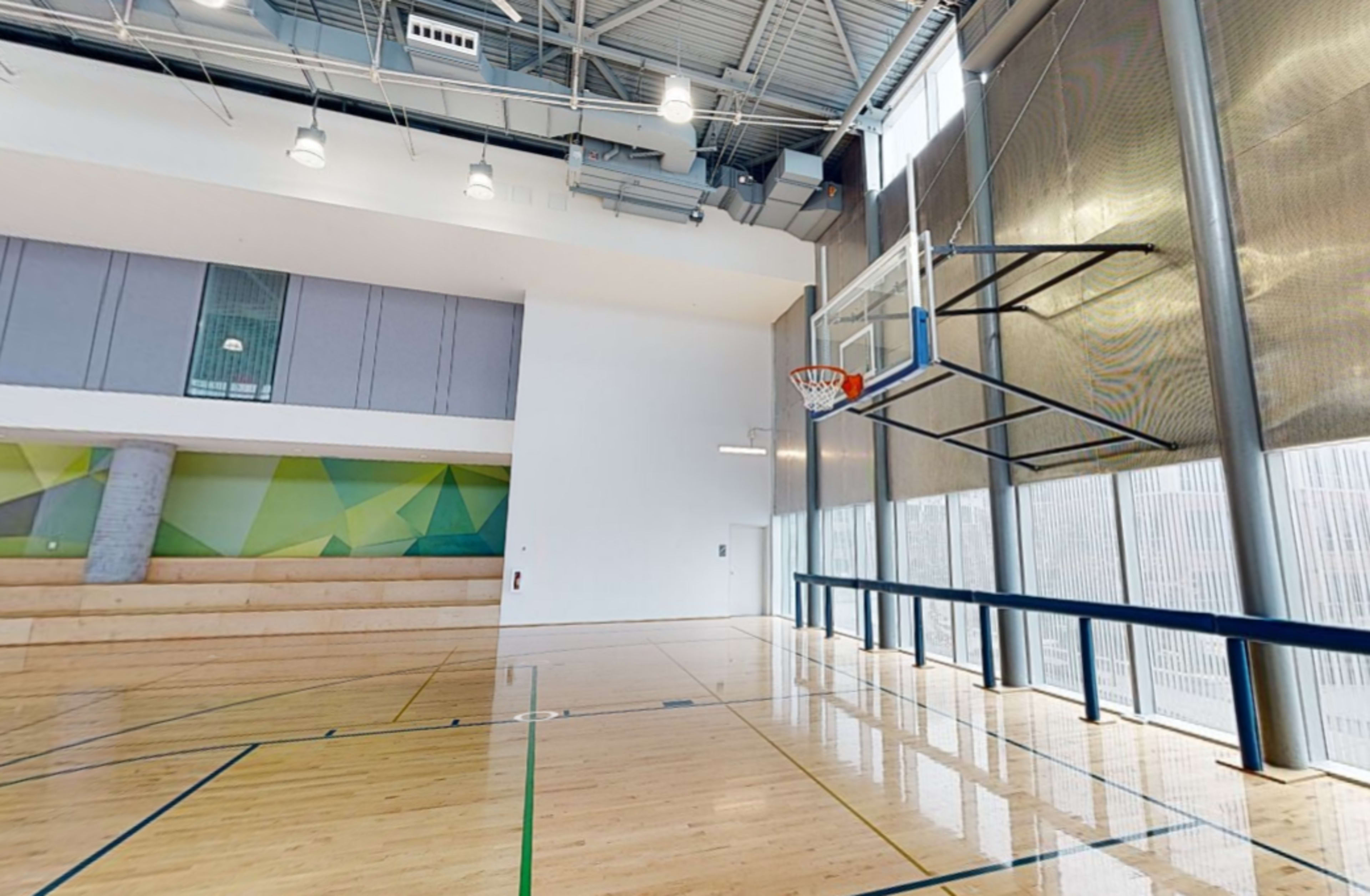 A spacious gymnasium with a basketball hoop mounted on a polished wooden floor and large windows allowing natural light to enter.