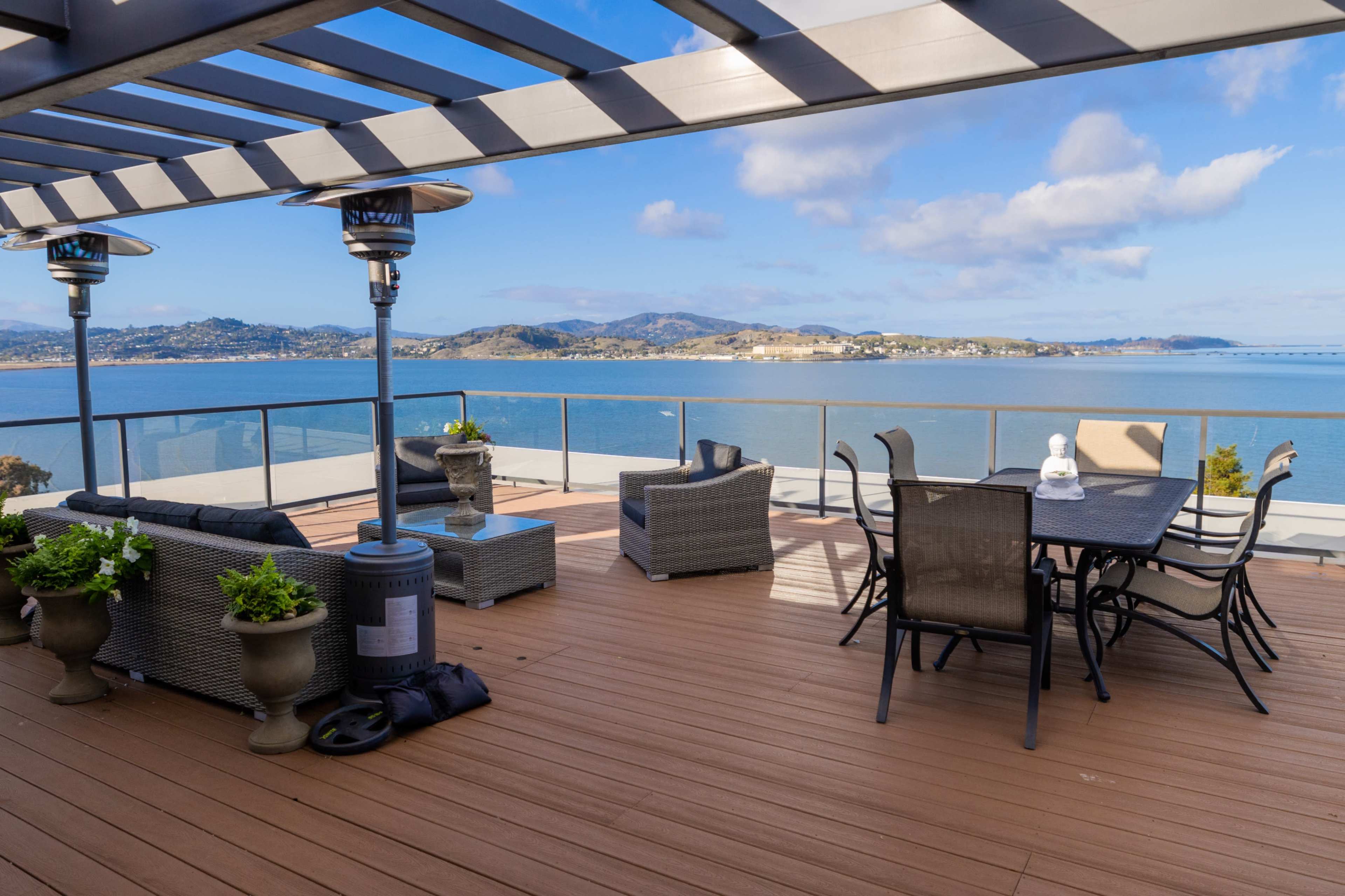 A spacious deck features outdoor furniture, heaters, and a panoramic view of a body of water and distant hills.