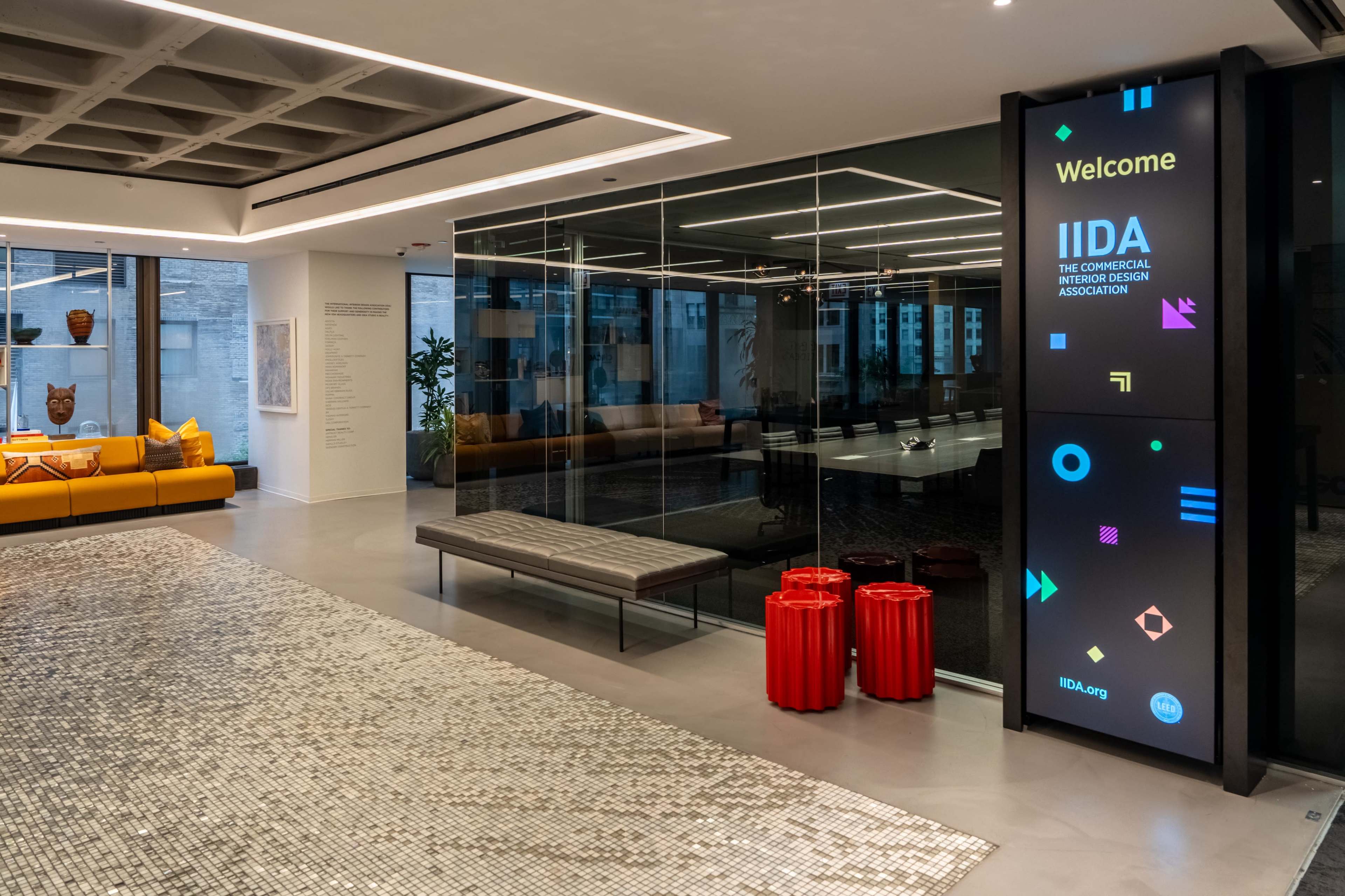 The image shows a modern office lobby featuring a digital welcome sign, a bench, red cylindrical stools, and a reflective floor, with large glass windows revealing a seating area behind.
