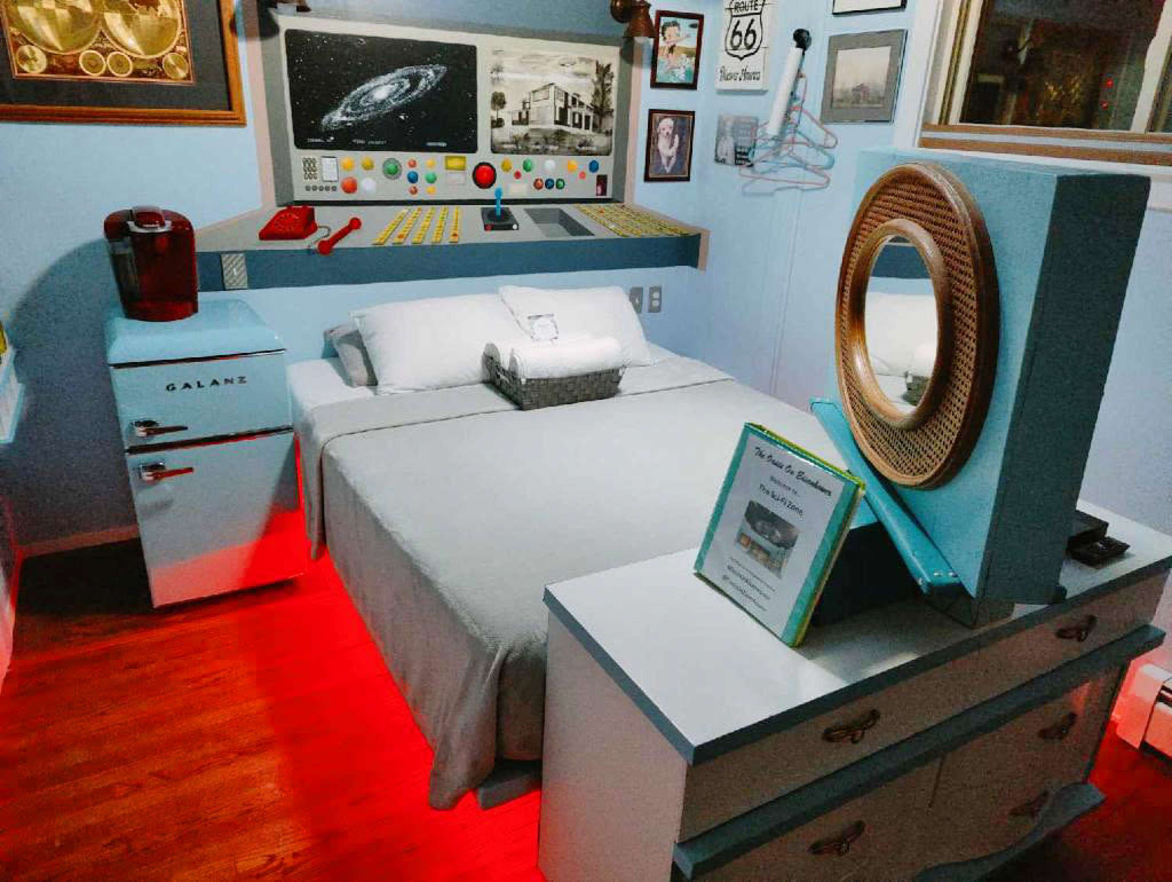 A creatively designed bedroom styled with vintage and retro elements, featuring a bed, a small fridge, and a vanity with a circular mirror against a light blue backdrop.