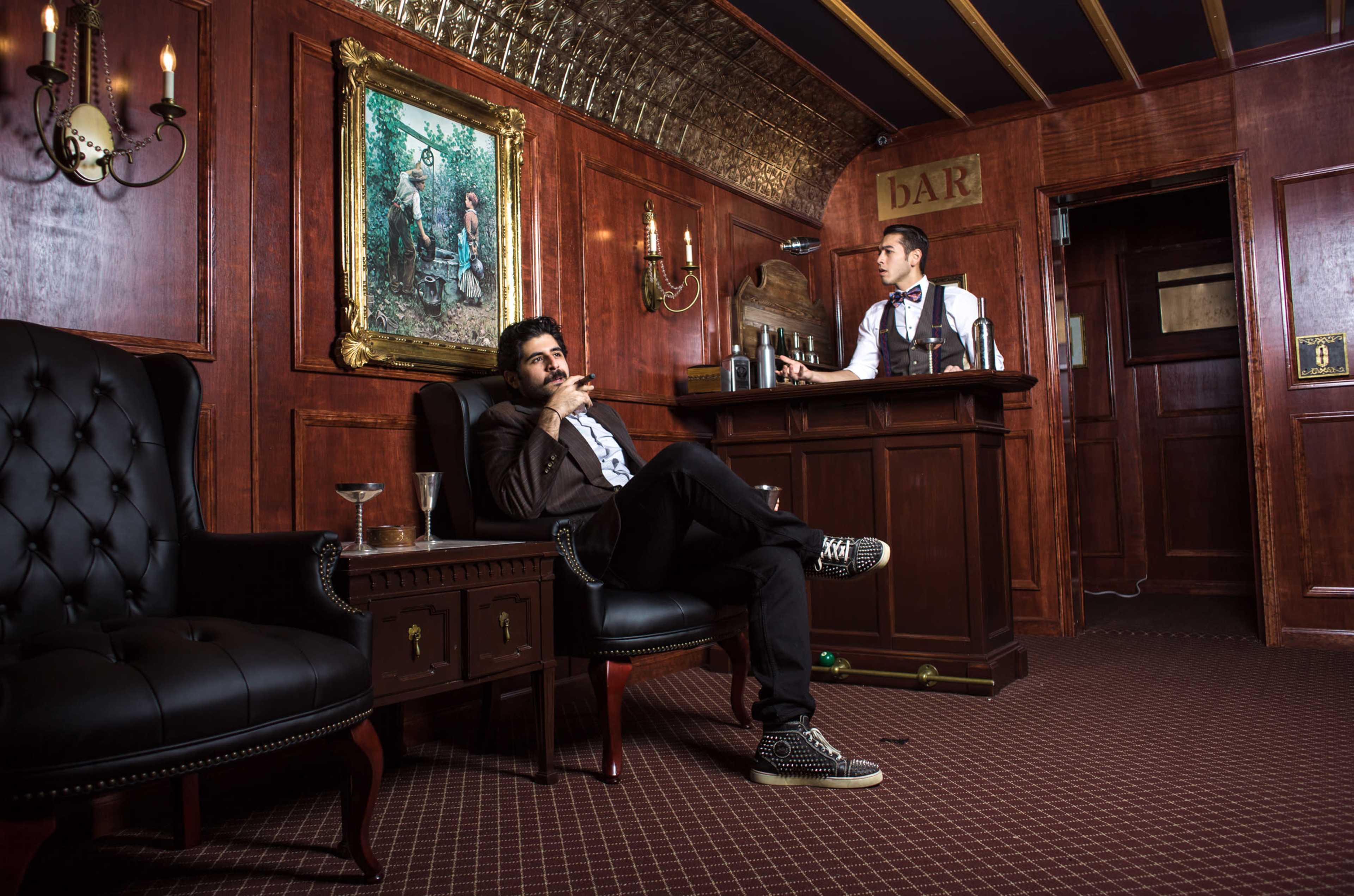 A man sits in an armchair smoking a cigar while another man stands behind a bar in a wooden-paneled room adorned with a large painting.