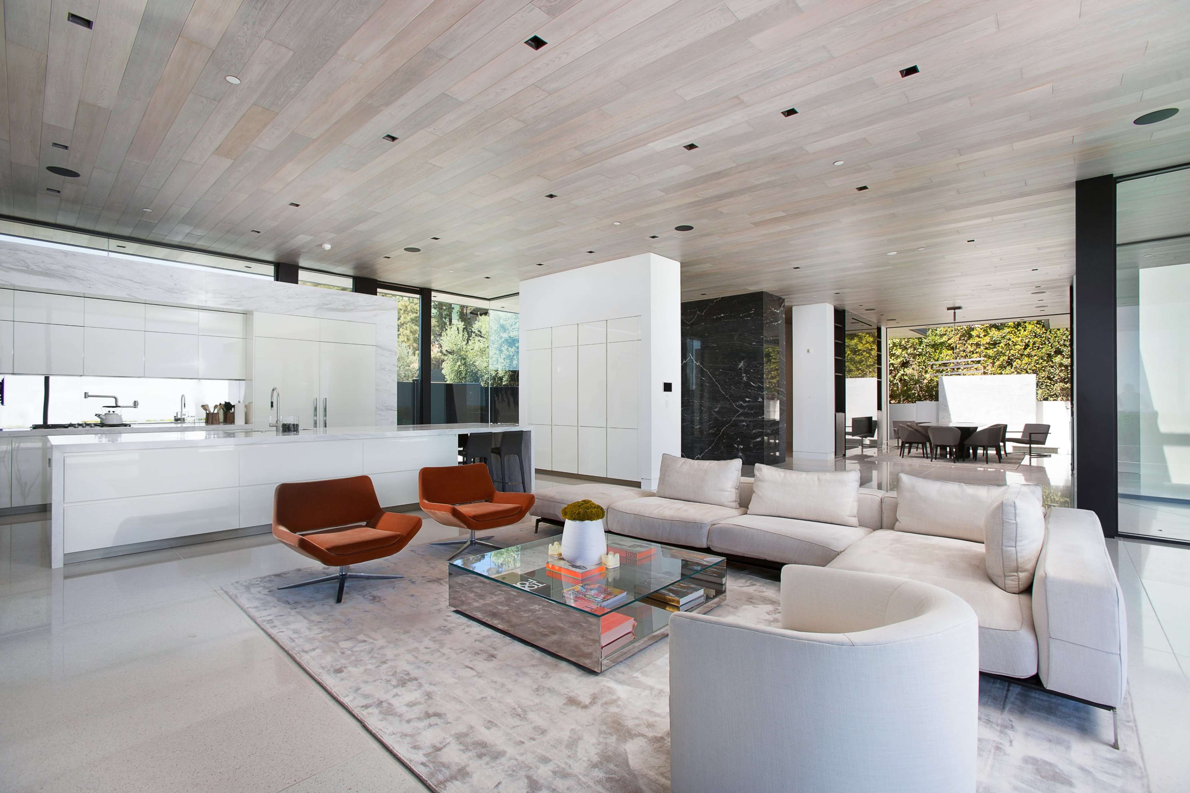 The image shows a modern living room with a large sectional sofa, two orange armchairs, a glass coffee table, and an open kitchen area with sleek cabinetry.