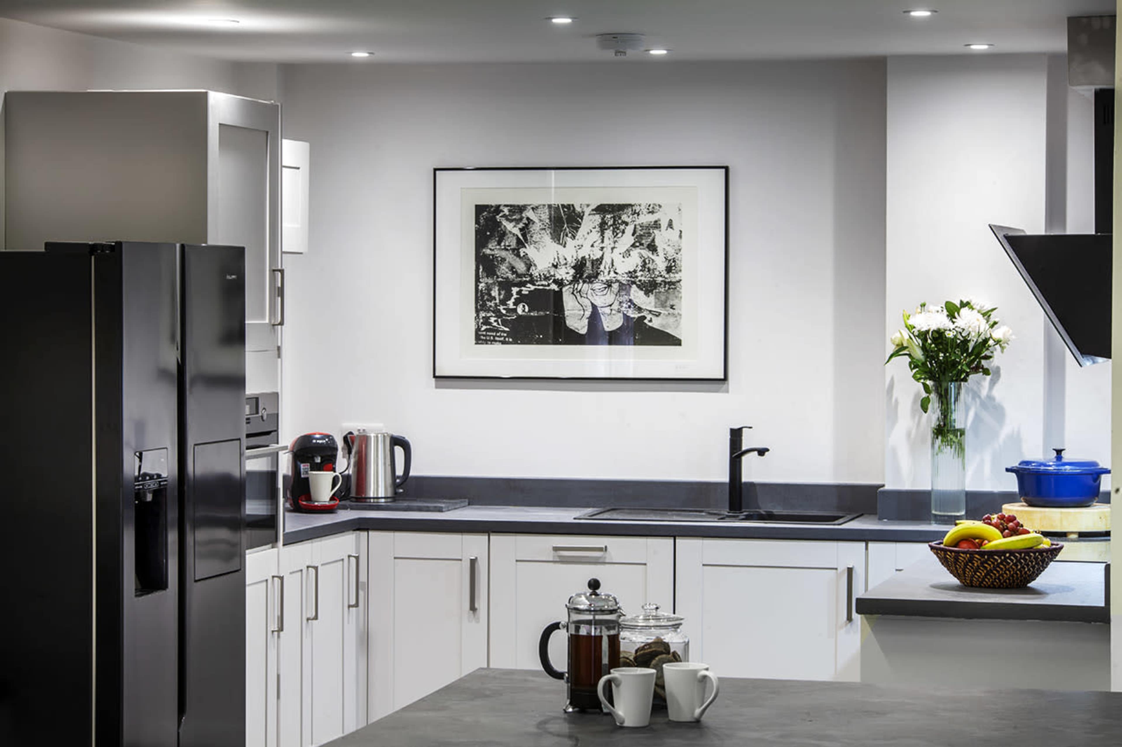 The image shows a modern kitchen with white cabinets, a black refrigerator, a countertop, a sink, and a variety of appliances including a coffee maker and a fruit bowl.