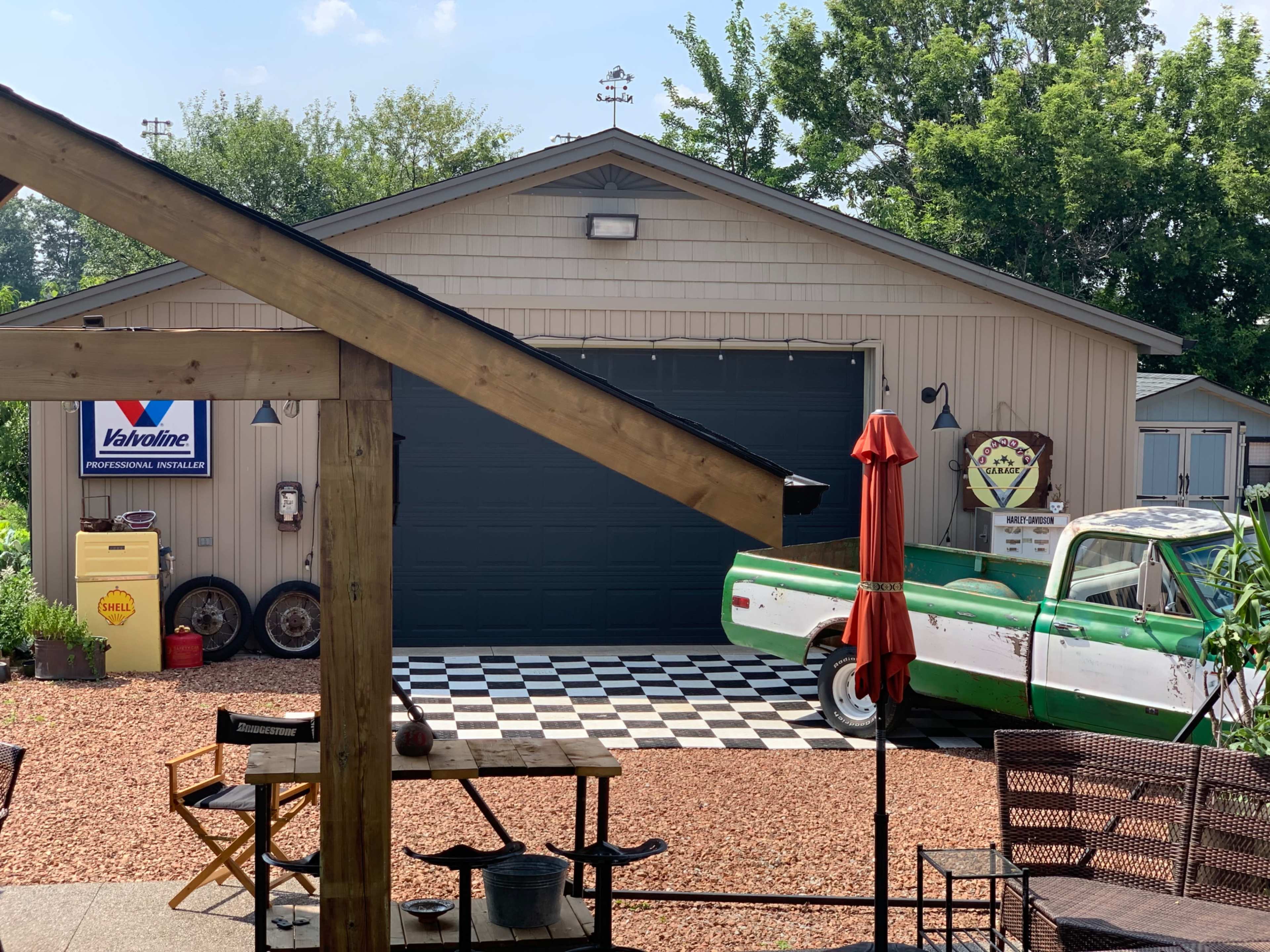 A vintage green and white pickup truck is parked in front of a garage with a checkered floor and various signs in a gravel yard.