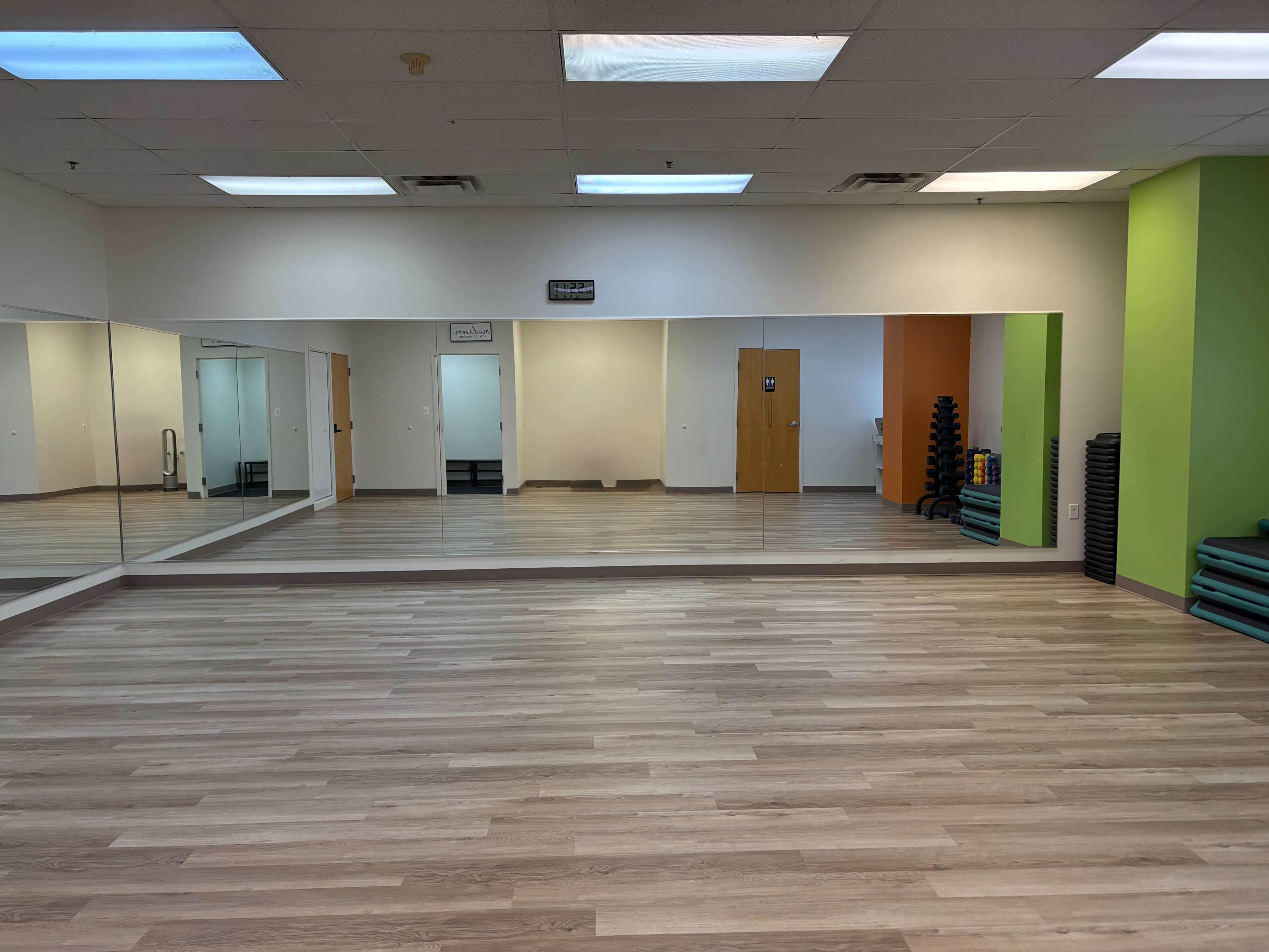 The image shows a bright, empty fitness studio with large mirrors on one wall, wooden flooring, and colorful walls.
