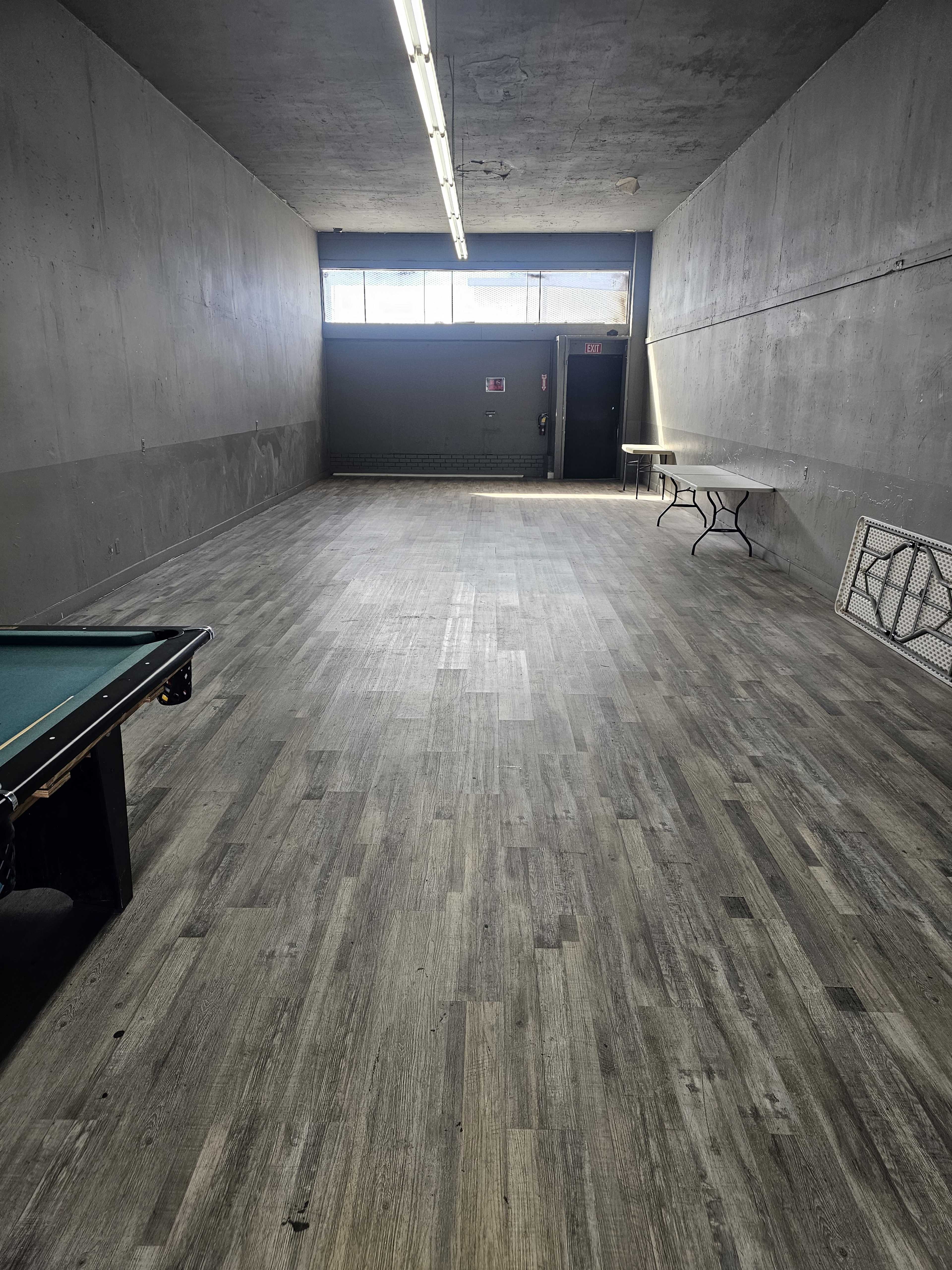 The image shows a large, empty room with gray walls, a wooden floor, and a pool table on the left side, along with several folding tables against the right wall.