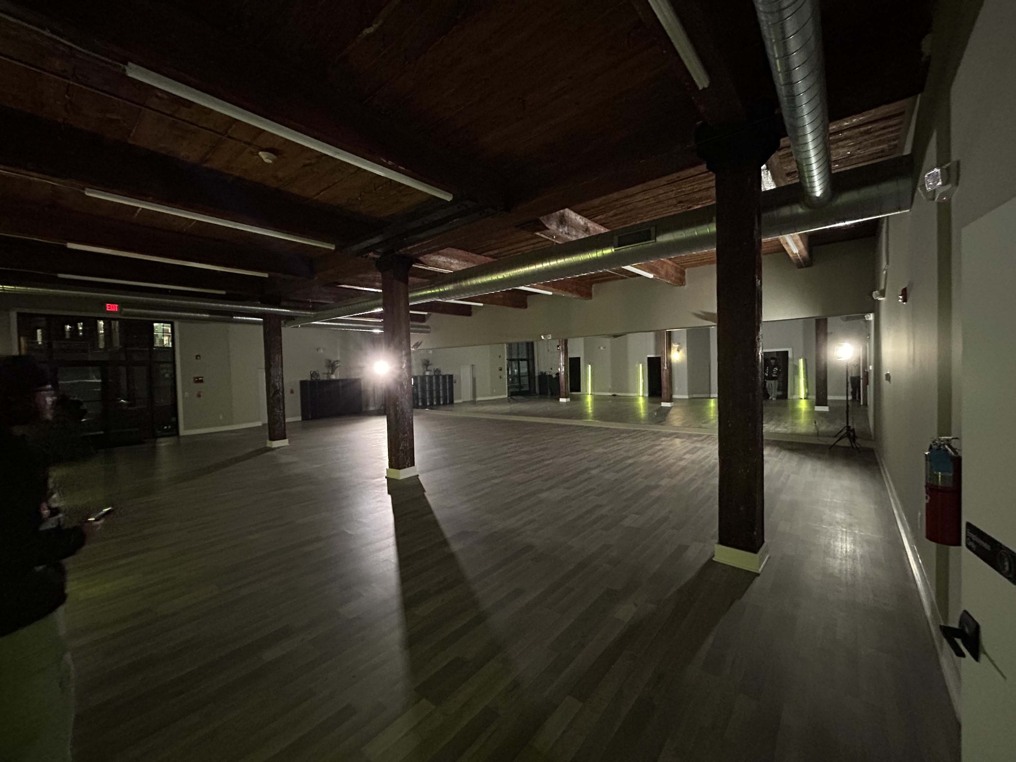 The image shows a spacious, dimly lit room with wooden beams and polished wooden flooring, featuring a minimal setup of lights and a bar area in the background.
