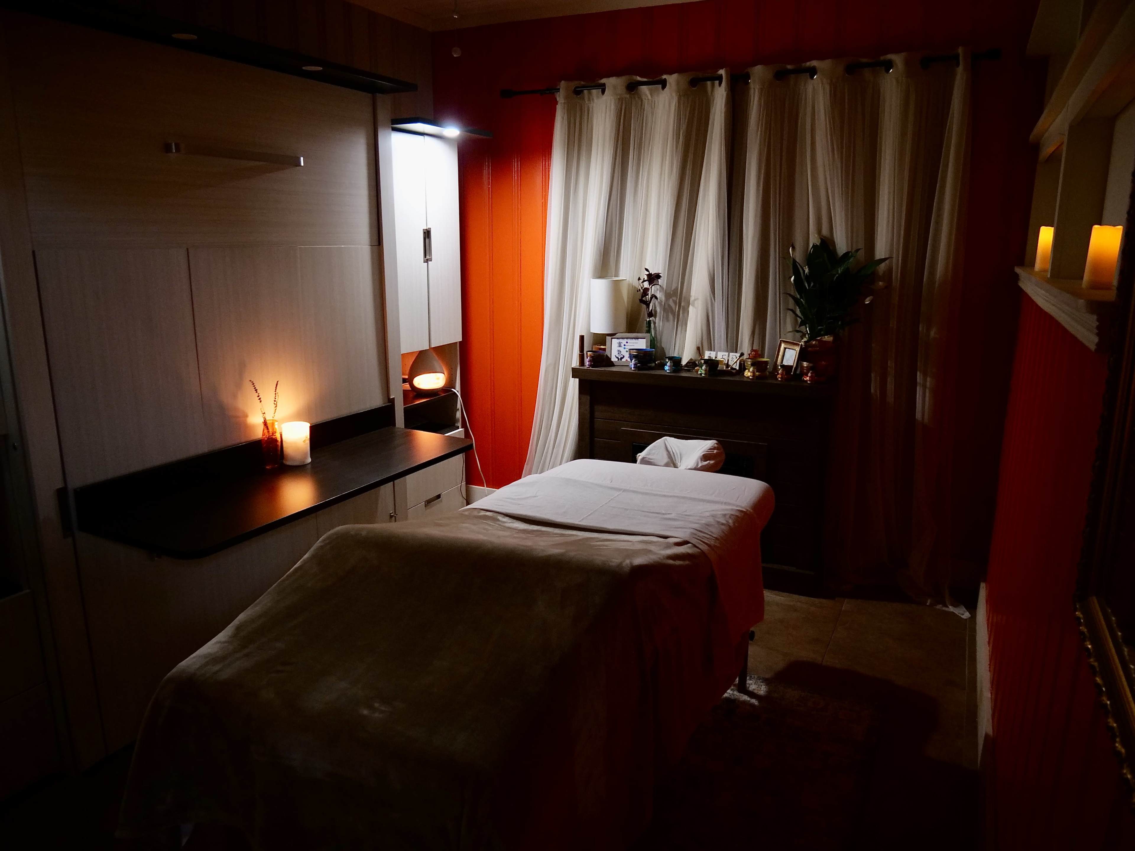 The image shows a softly lit spa room with a massage table, surrounded by candles, curtains, and decorative items on a dresser.