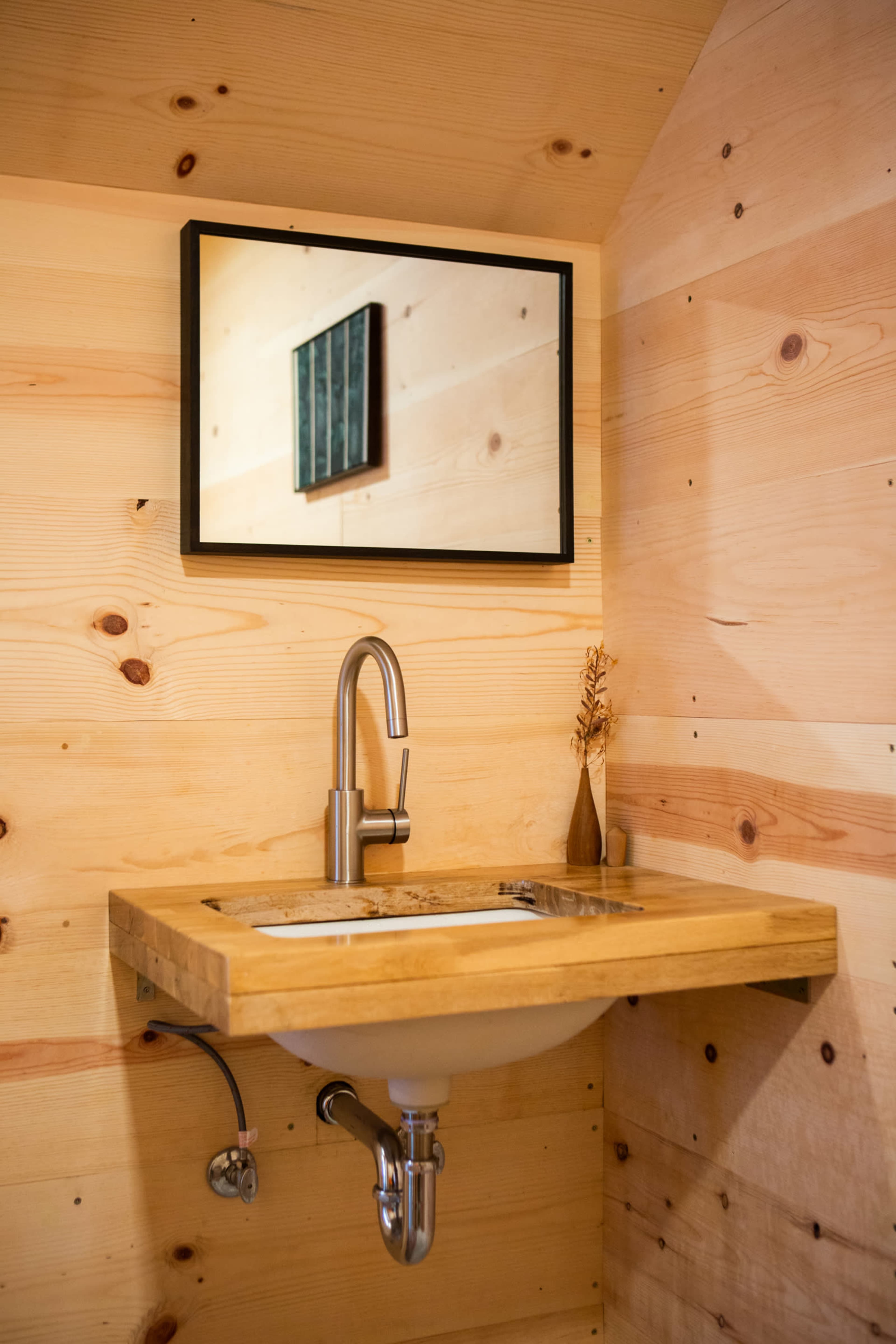 A wooden sink with a minimalist faucet is mounted on a wall with light-colored wood paneling, reflecting a simple framed artwork in the mirror above it.