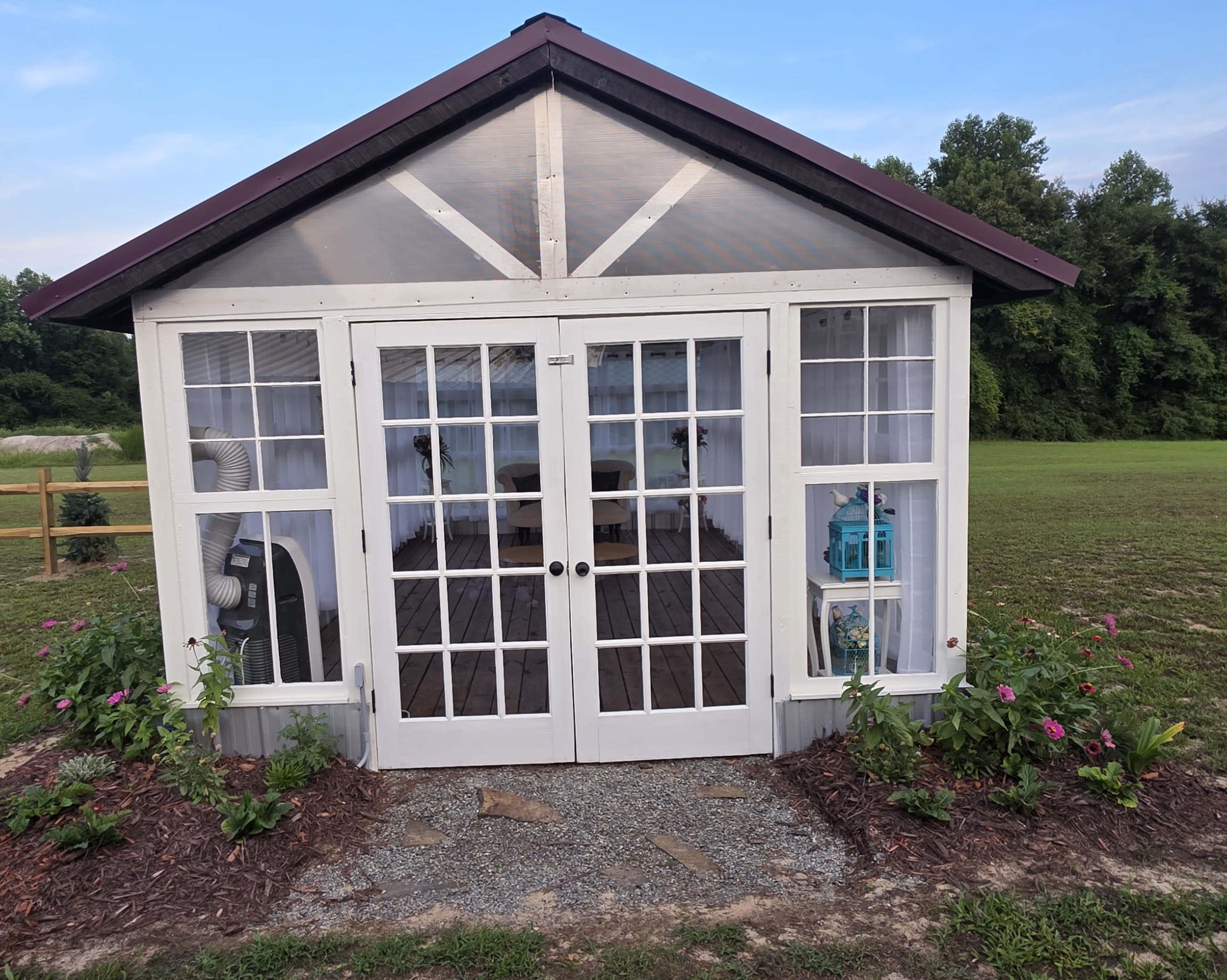 A small glass-enclosed building with double doors and a peaked roof is surrounded by flower beds and located on a grassy field.