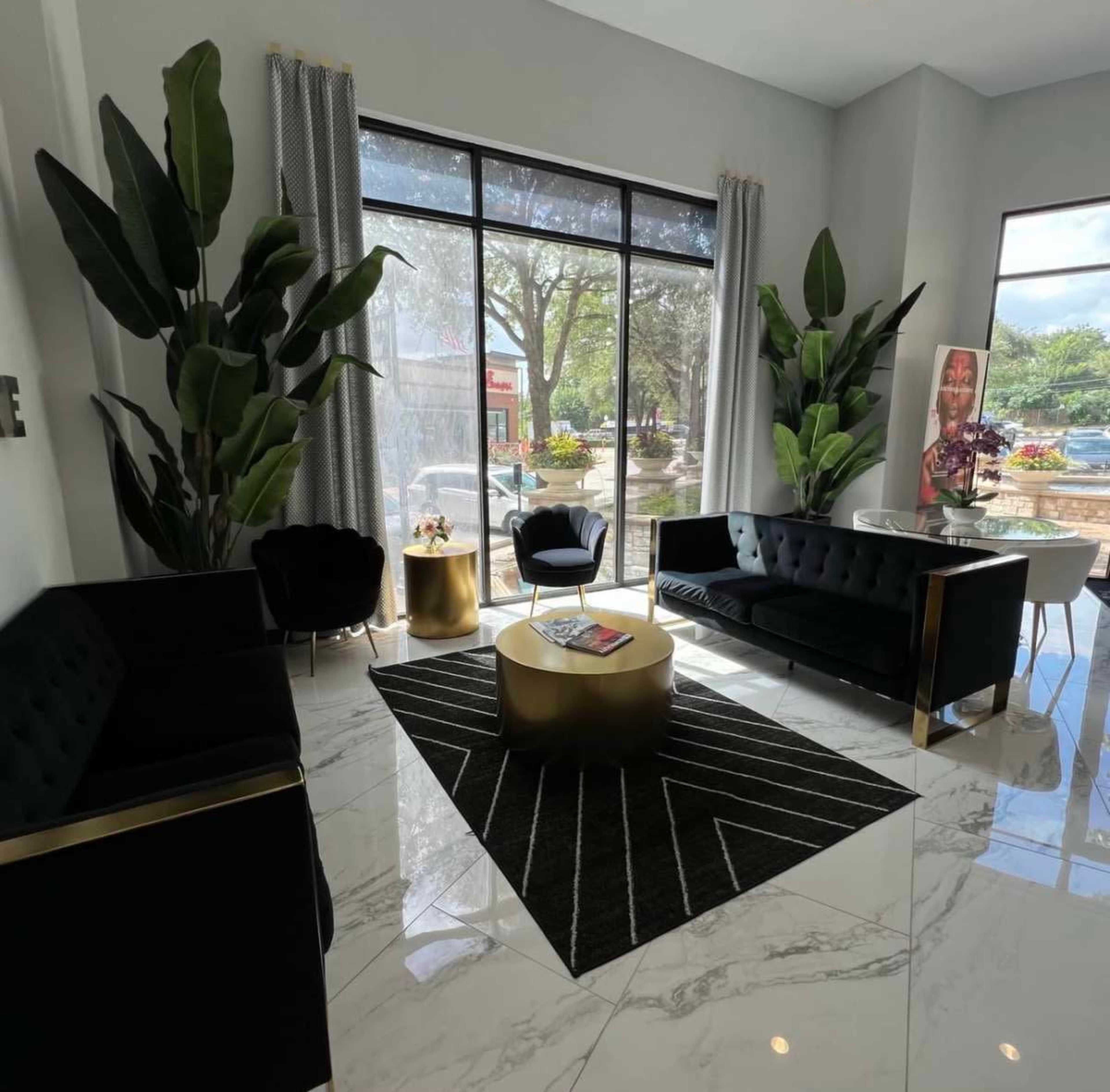 The image shows a modern seating area with black sofas, a round gold coffee table, and large windows featuring indoor plants and natural light.