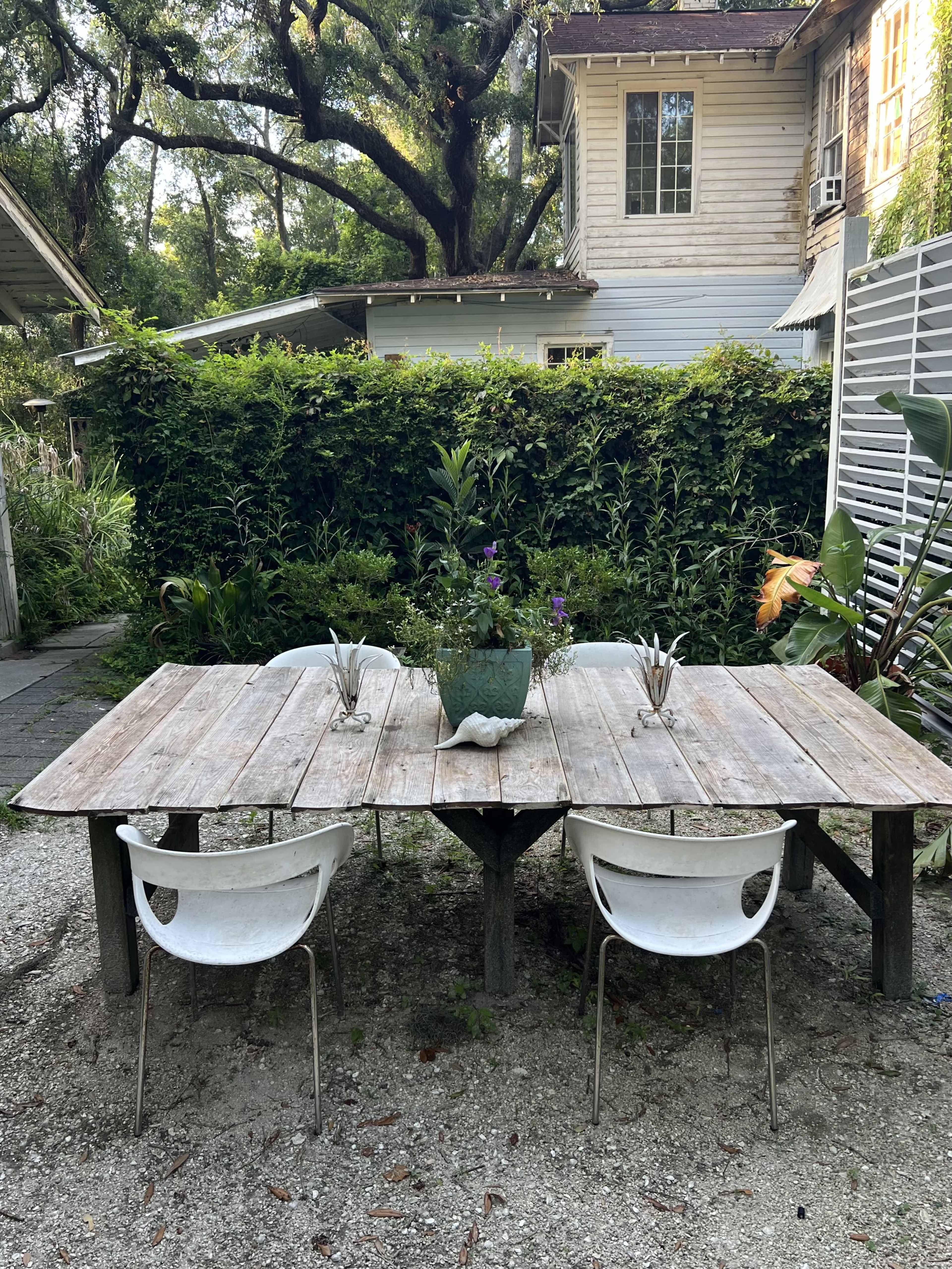 A weathered wooden table with two white chairs is set in an outdoor area surrounded by greenery and a house in the background.