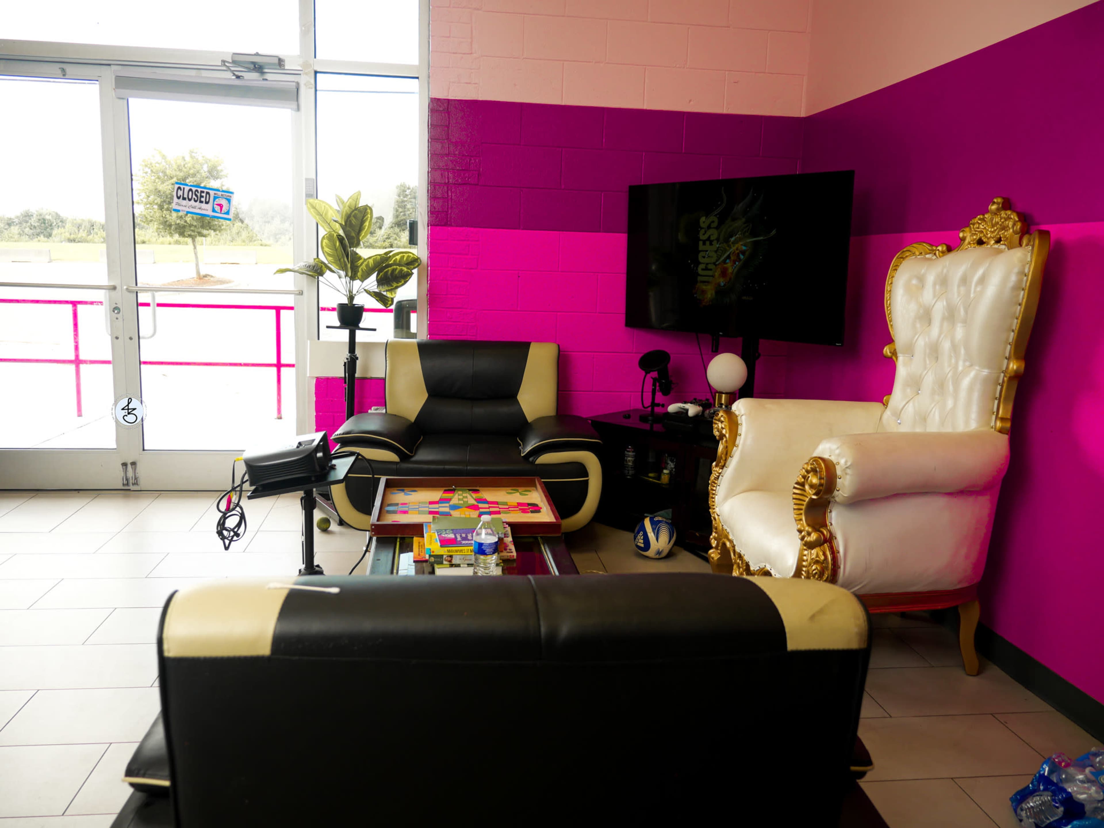 The image shows a brightly colored seating area with black and white chairs, a large ornate white chair, a coffee table with games, and a television against a pink wall.