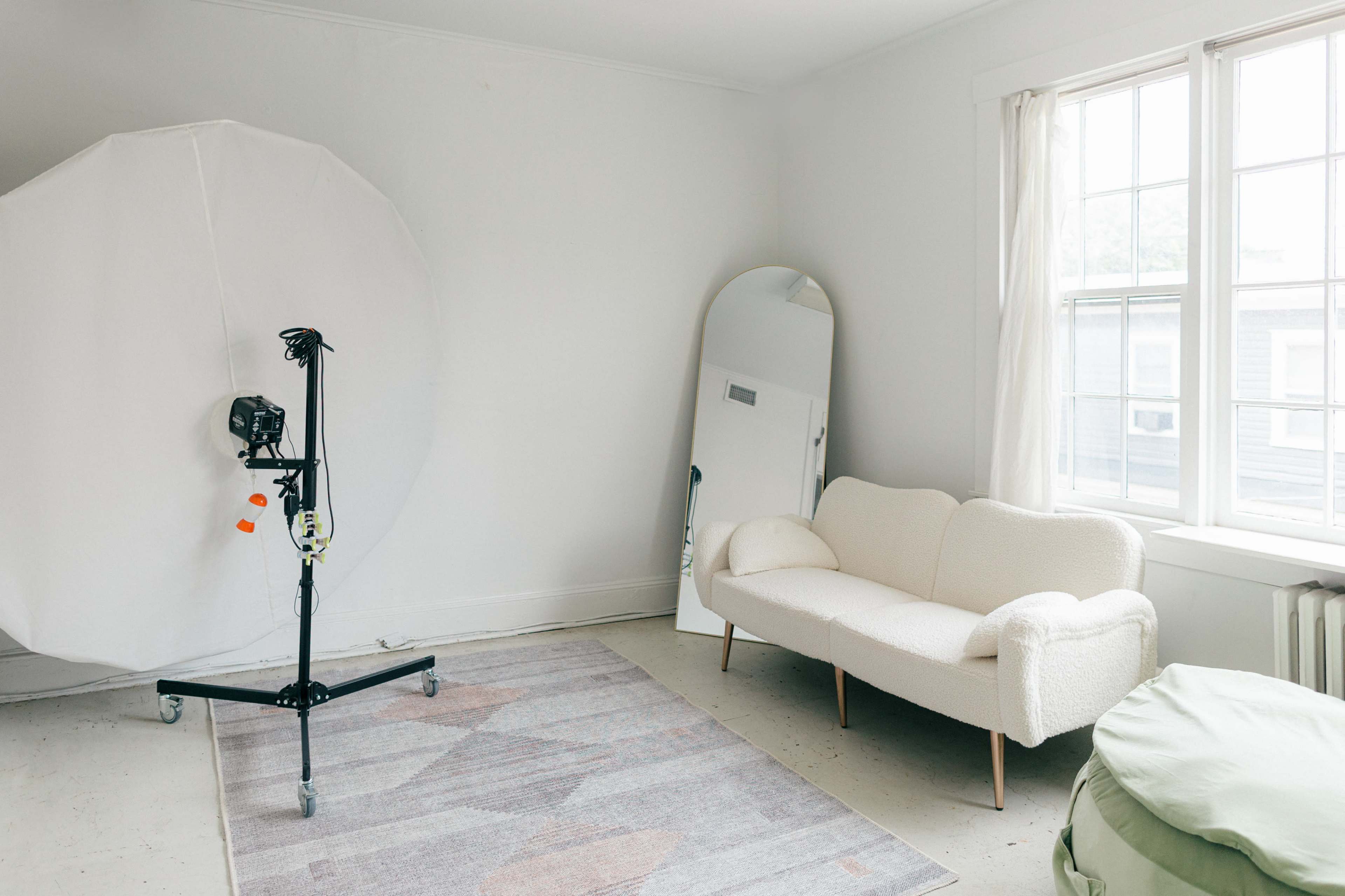 The image shows a minimalistic room with a white couch, a standing mirror, a large softbox light on a stand, and a patterned rug on the floor.