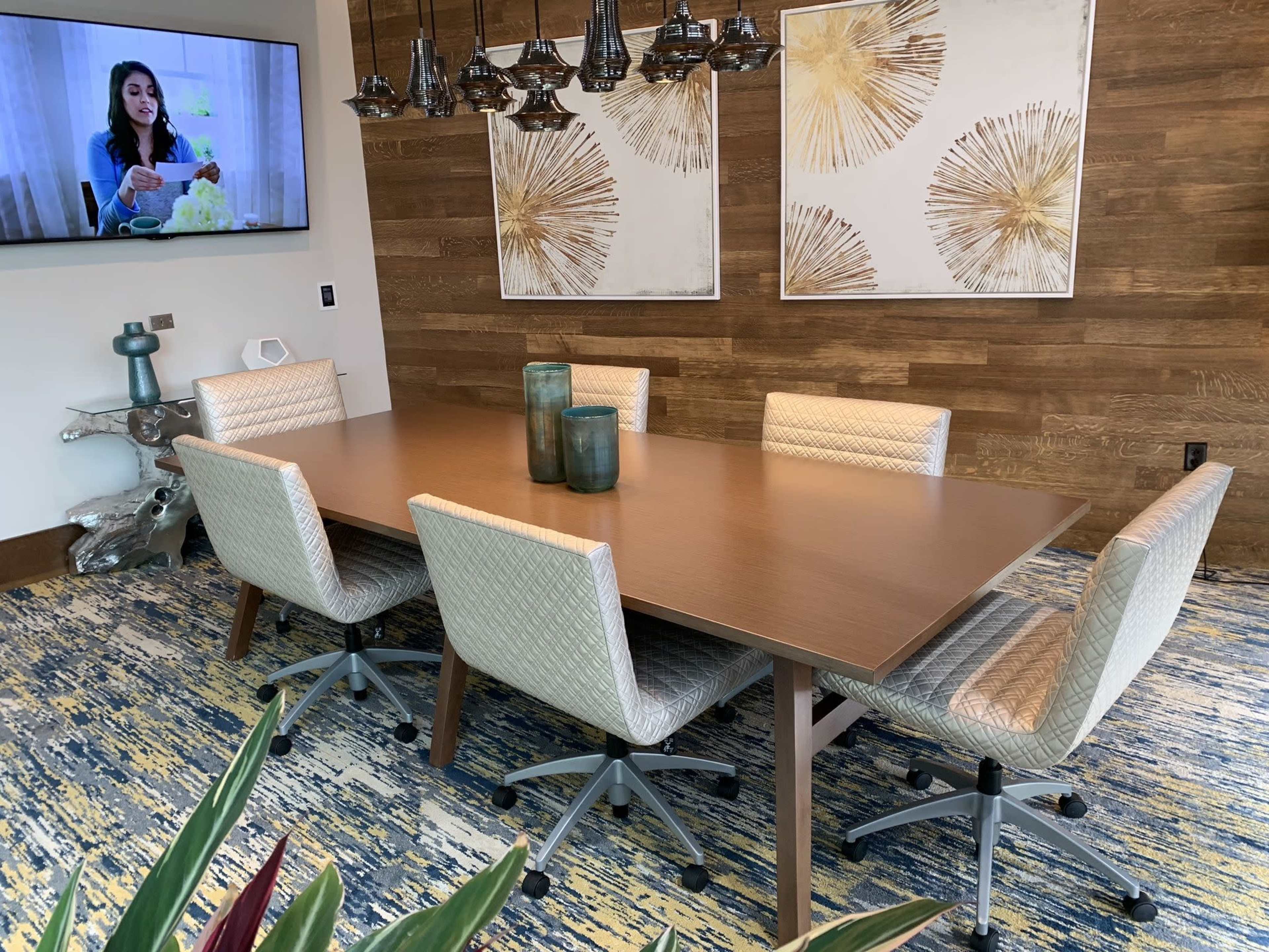 A dining area features a large wooden table surrounded by six upholstered chairs, with a television displaying a woman and decorative artwork on the walls.